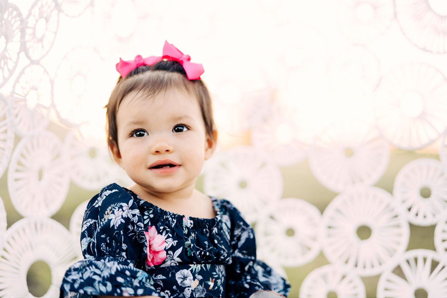A small child wearing a navy floral dress and pink bow smiles sweetly against a white lace backdrop.