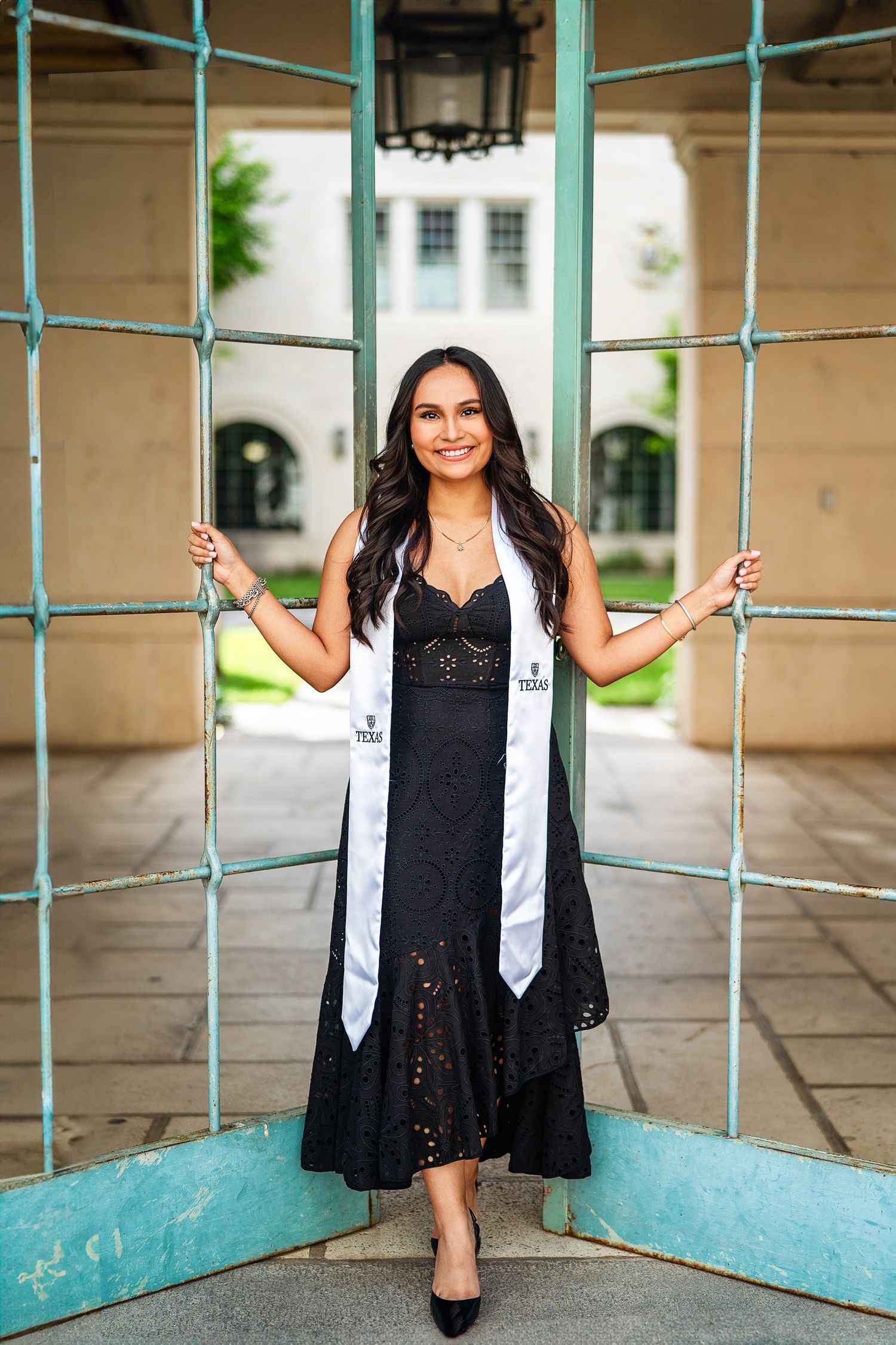 Graduate in black lace dress and white graduation stole poses by turquoise railings in an elegant archway.