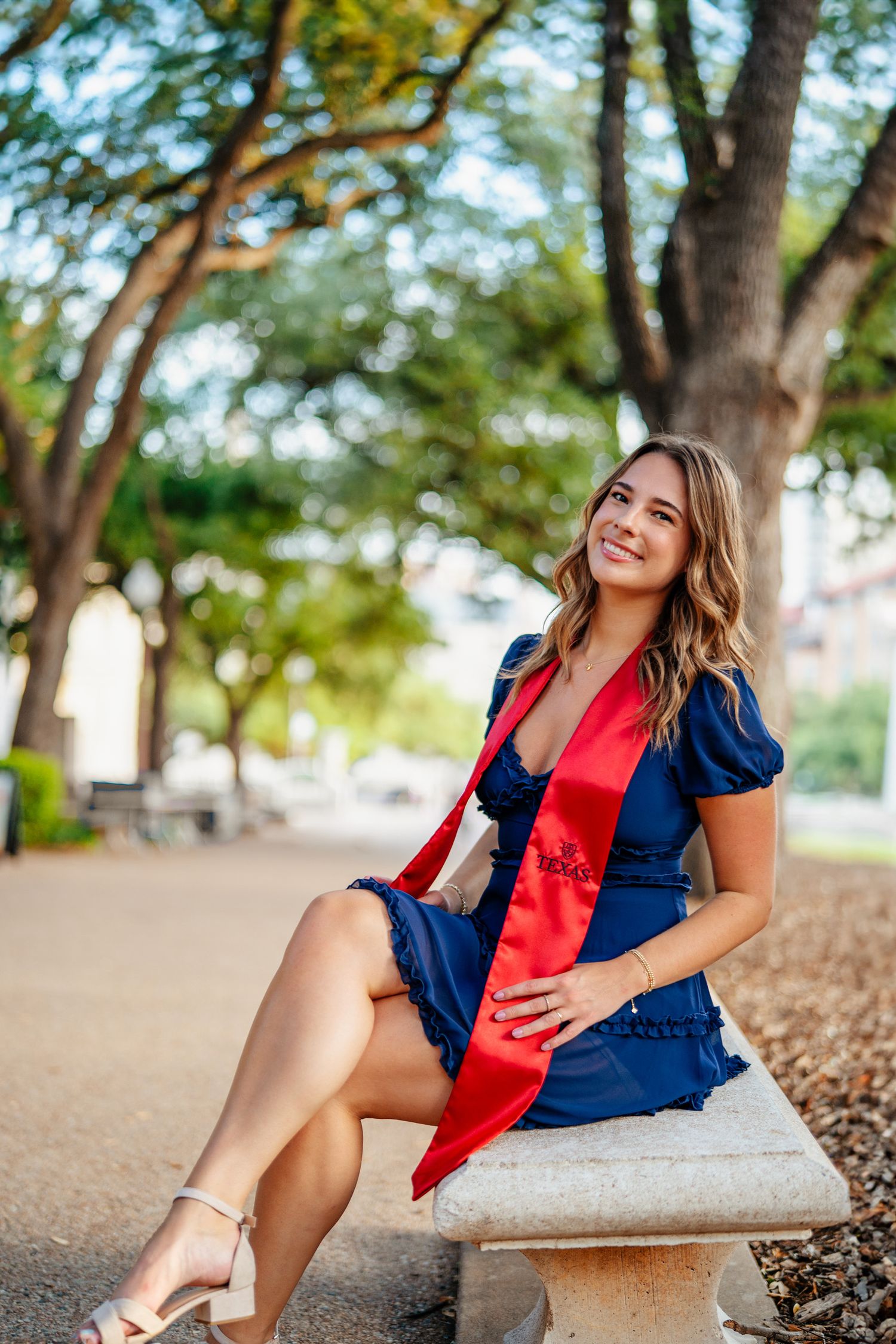 Graduate in navy dress and red stole poses on stone bench under trees at university campus.