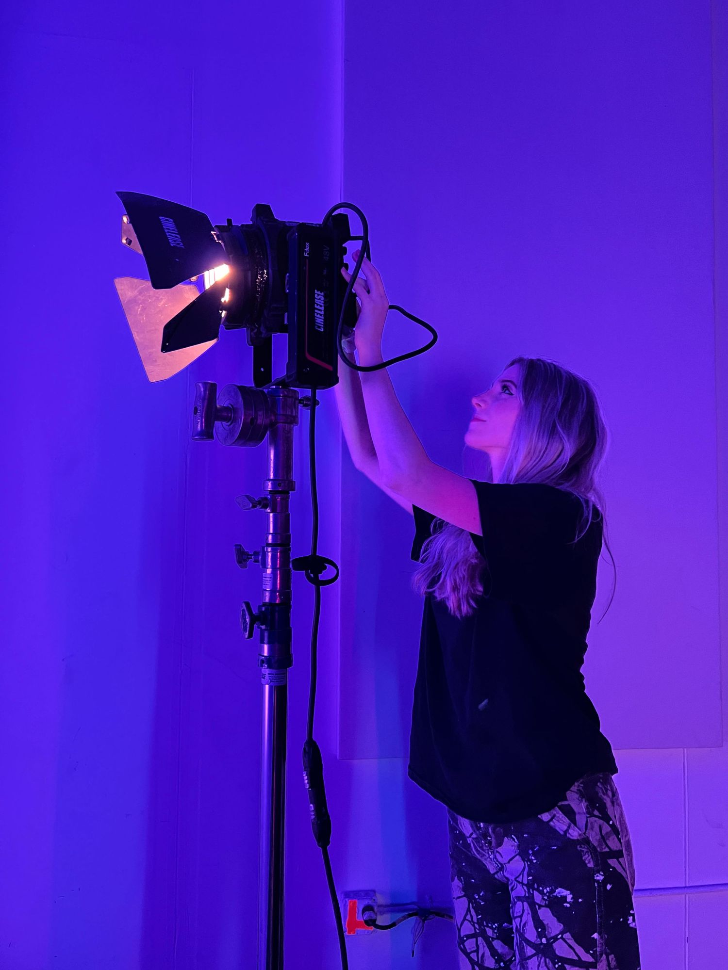 A crew member adjusts professional lighting equipment against a purple-lit background.