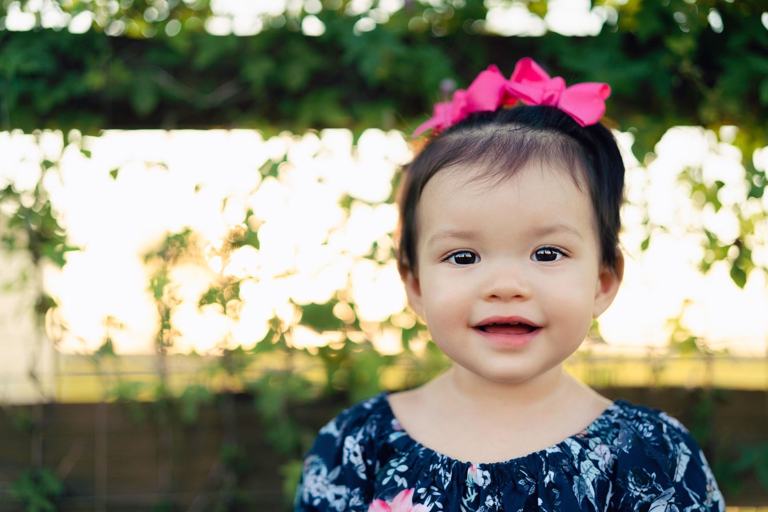 A joyful child in a navy dress with pink hair bow smiles outdoors in natural lighting.