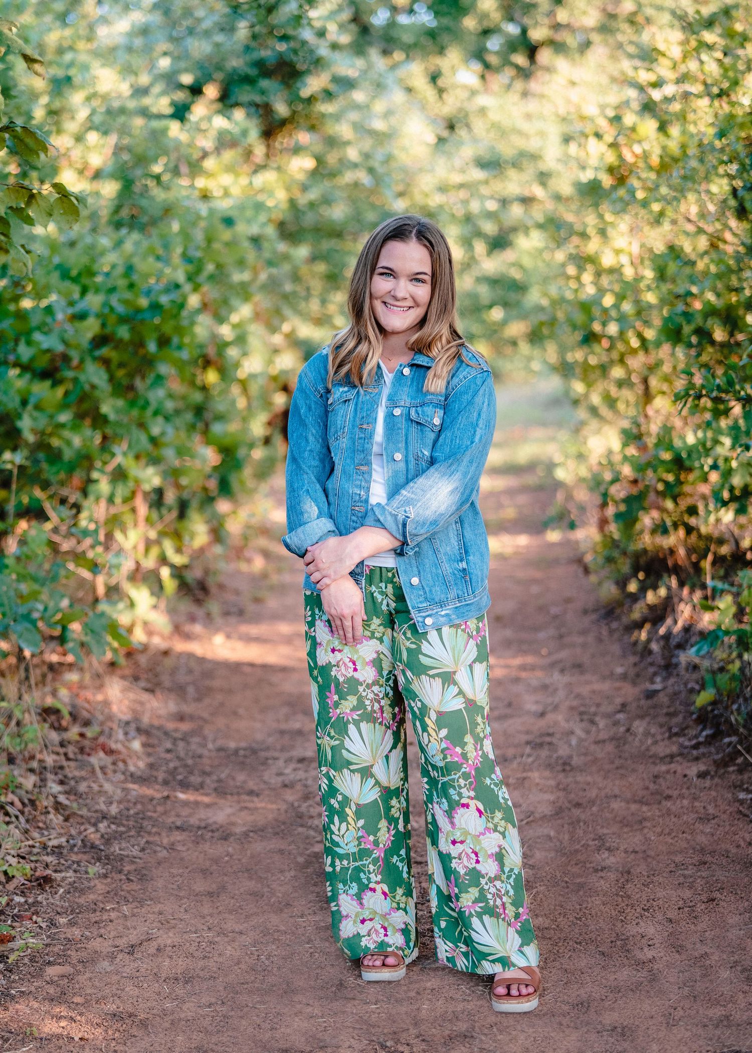 Person wearing denim jacket and floral pants poses on dirt path surrounded by greenery.