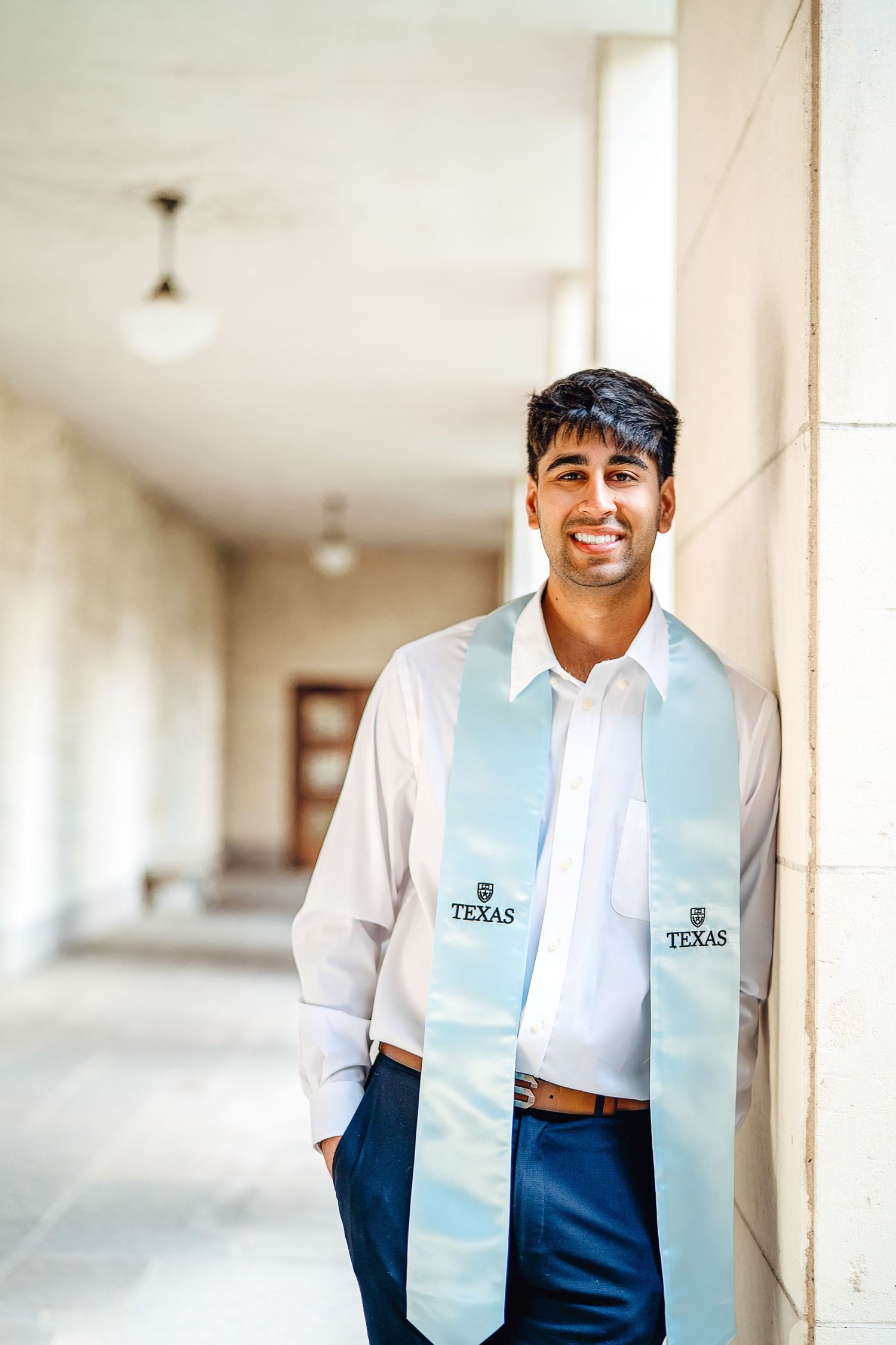 Graduate wearing light blue graduation stole with Texas logo posing in university hallway.