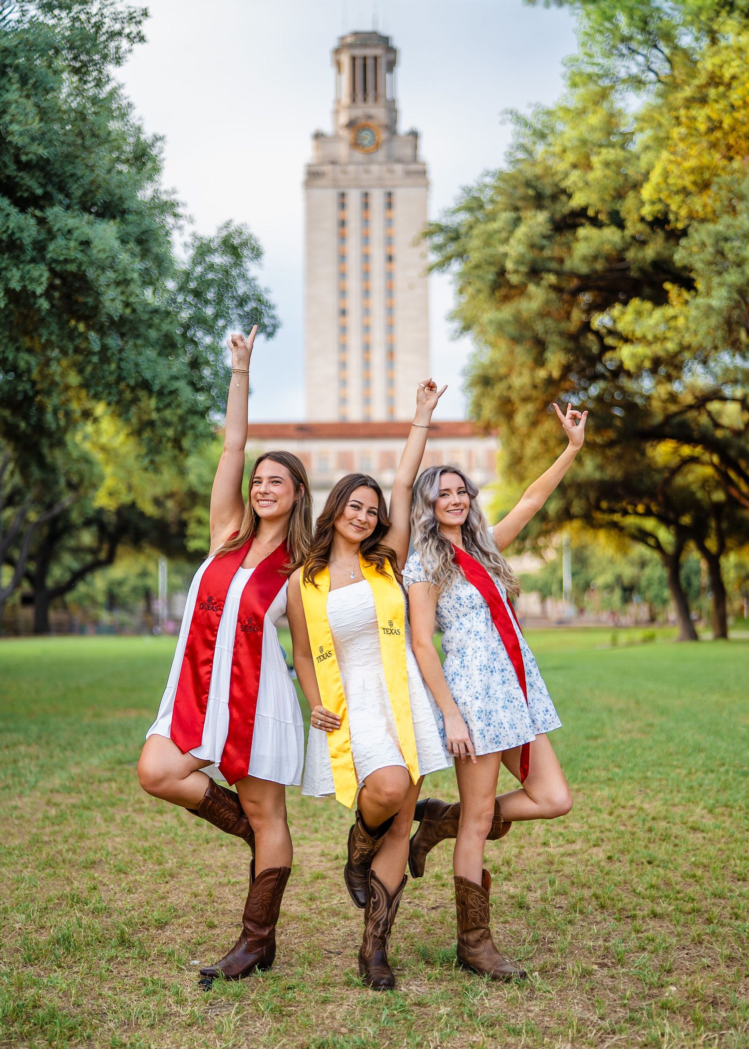 Three graduates in colorful dresses and cowboy boots celebrate in front of UT Austin tower.