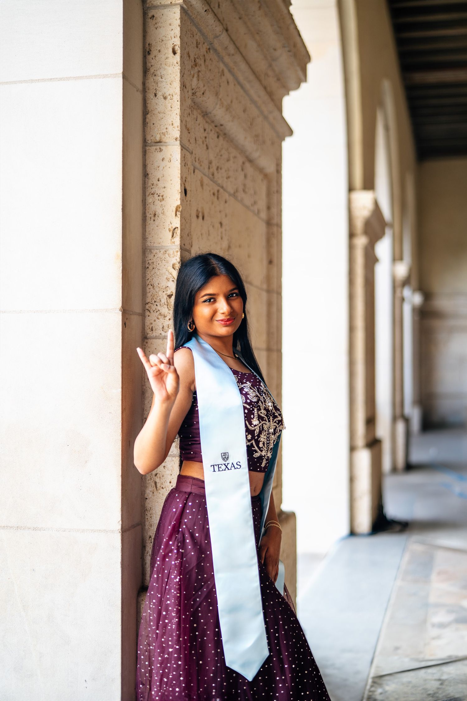 Graduate in purple gown and white stole poses in columned university corridor.
