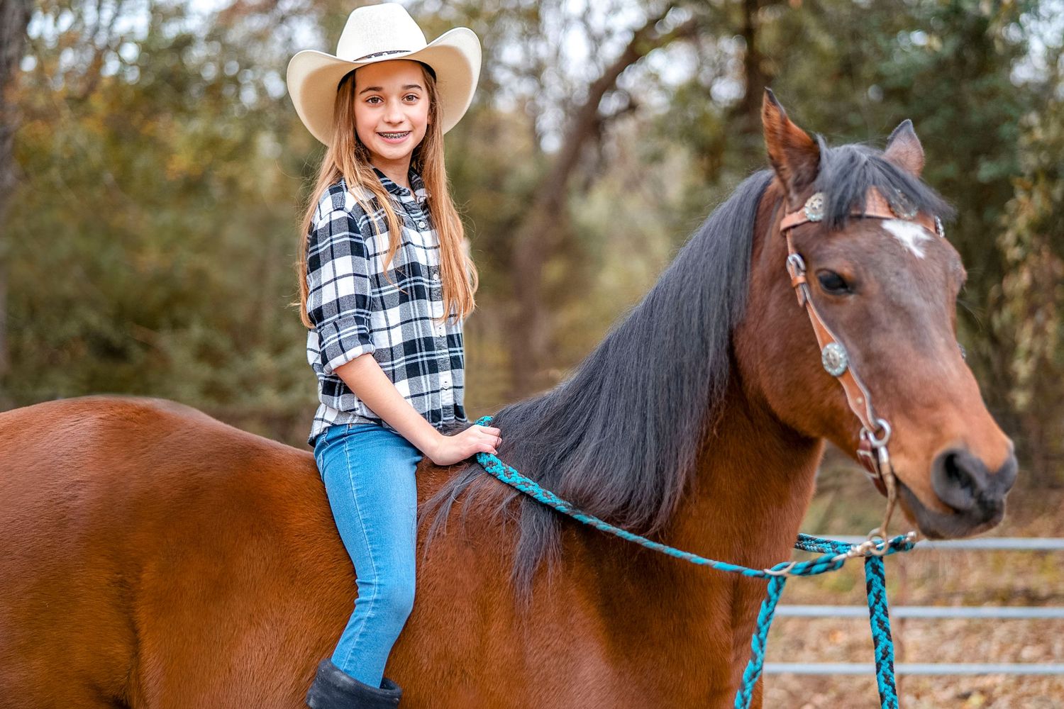 Person in cowboy hat and plaid shirt riding a brown horse in an outdoor setting.