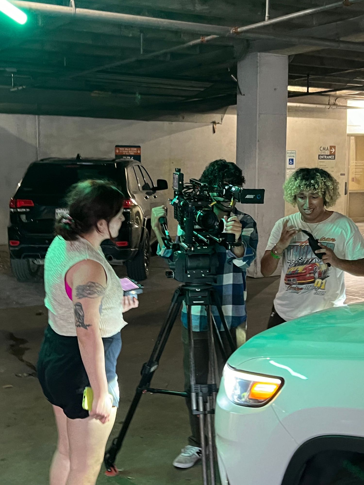 Film crew sets up camera equipment in a parking garage with green lighting.