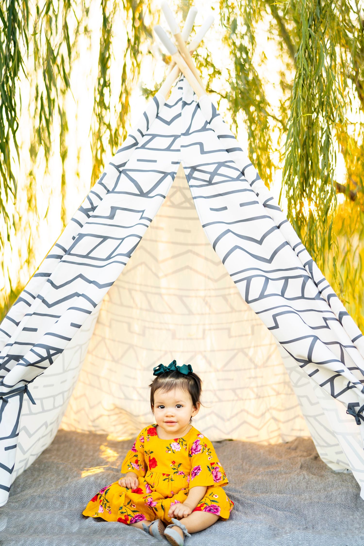 A child in a yellow floral dress sits in front of a white and blue striped teepee tent.