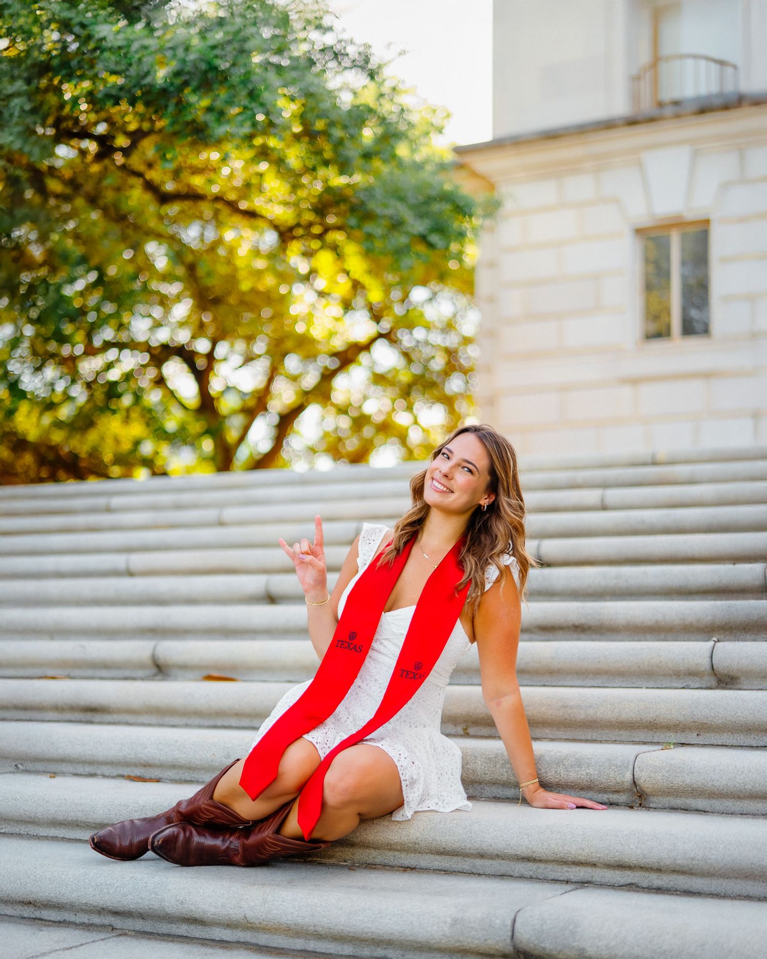 A woman in a red and white dress sits on stone steps near autumn foliage and a building.