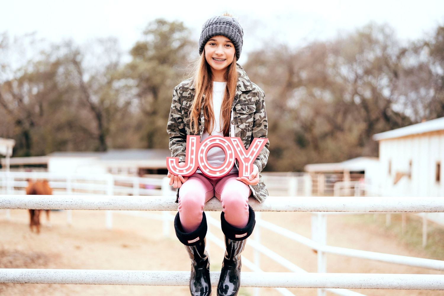 Person in winter hat and pink clothing poses near white fencing on a farm.