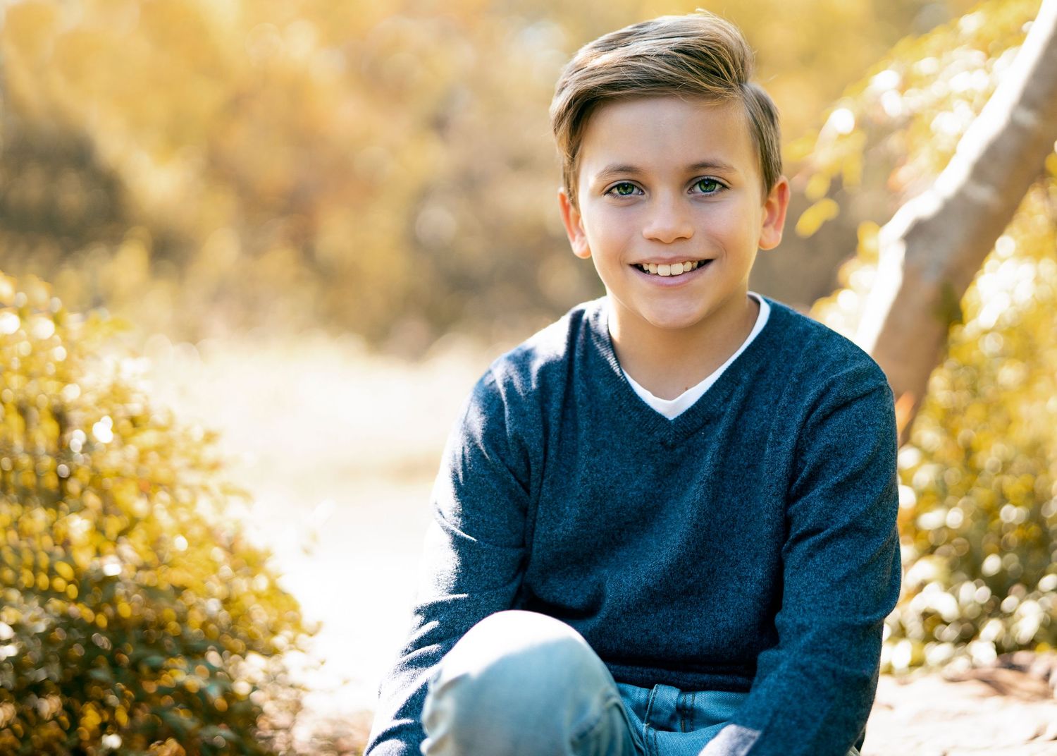 Young person in navy sweater sitting outdoors with golden autumn foliage in background.