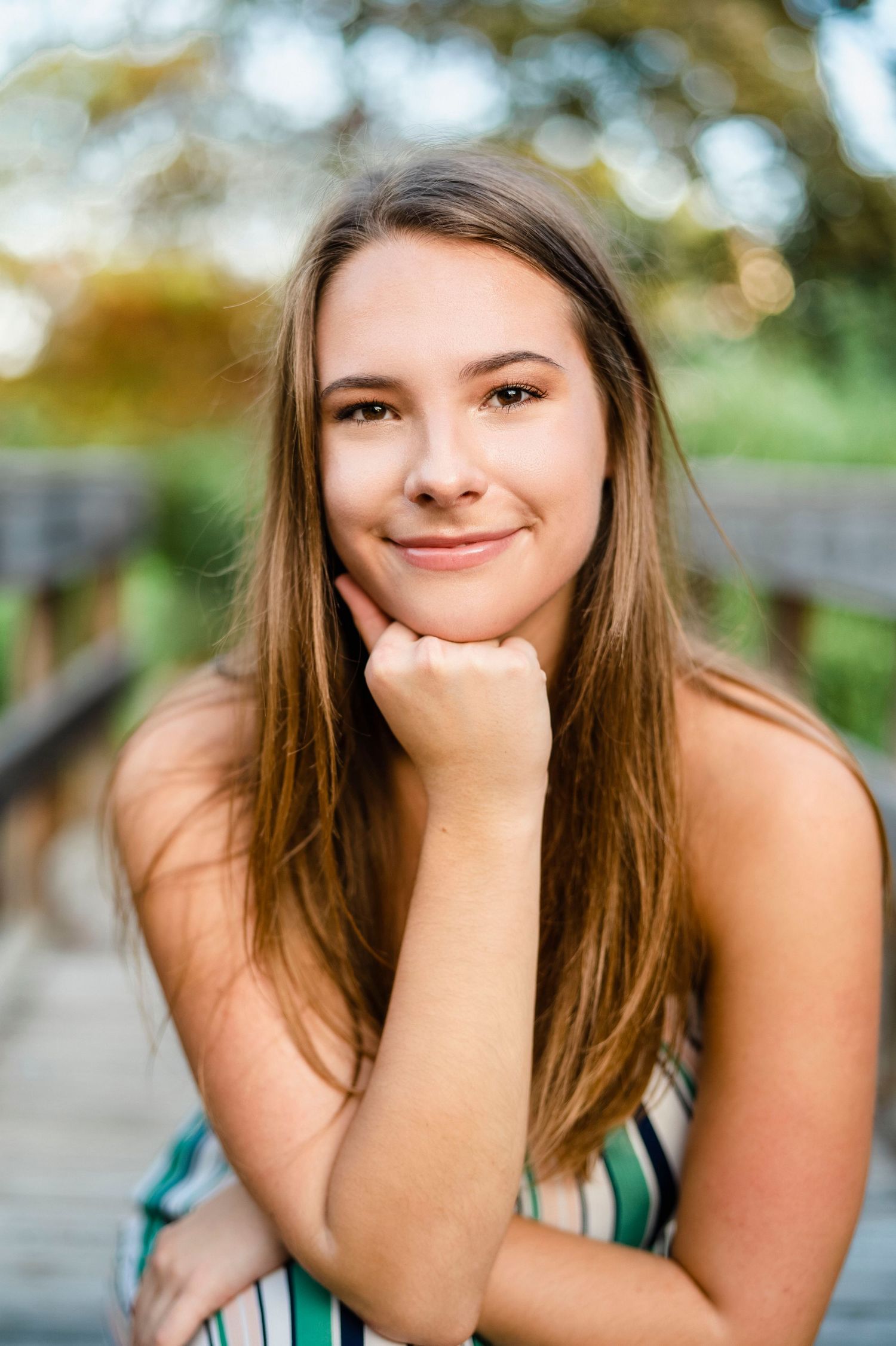 Portrait of a smiling woman with long brown hair resting her chin on her hand outdoors.