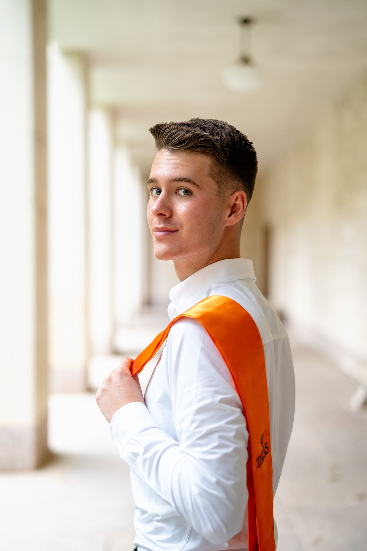 Graduate in white shirt and orange stole poses in hallway during graduation photos.