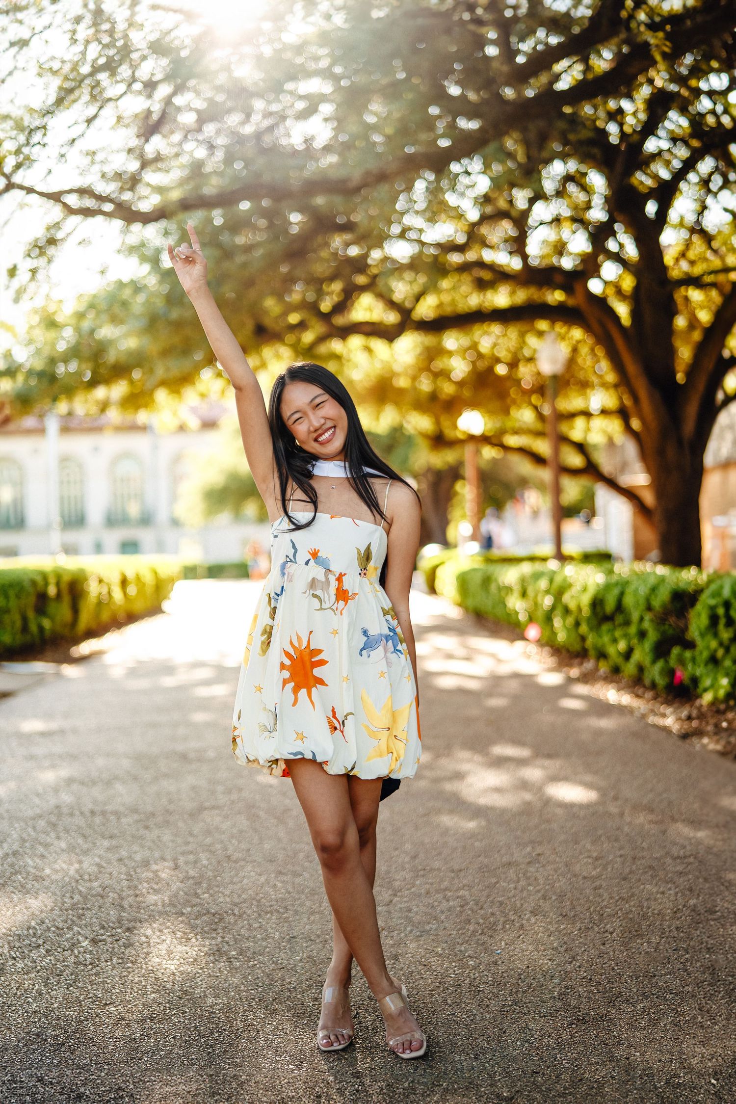 Graduate in white floral sundress poses joyfully on campus pathway during golden hour sunset.