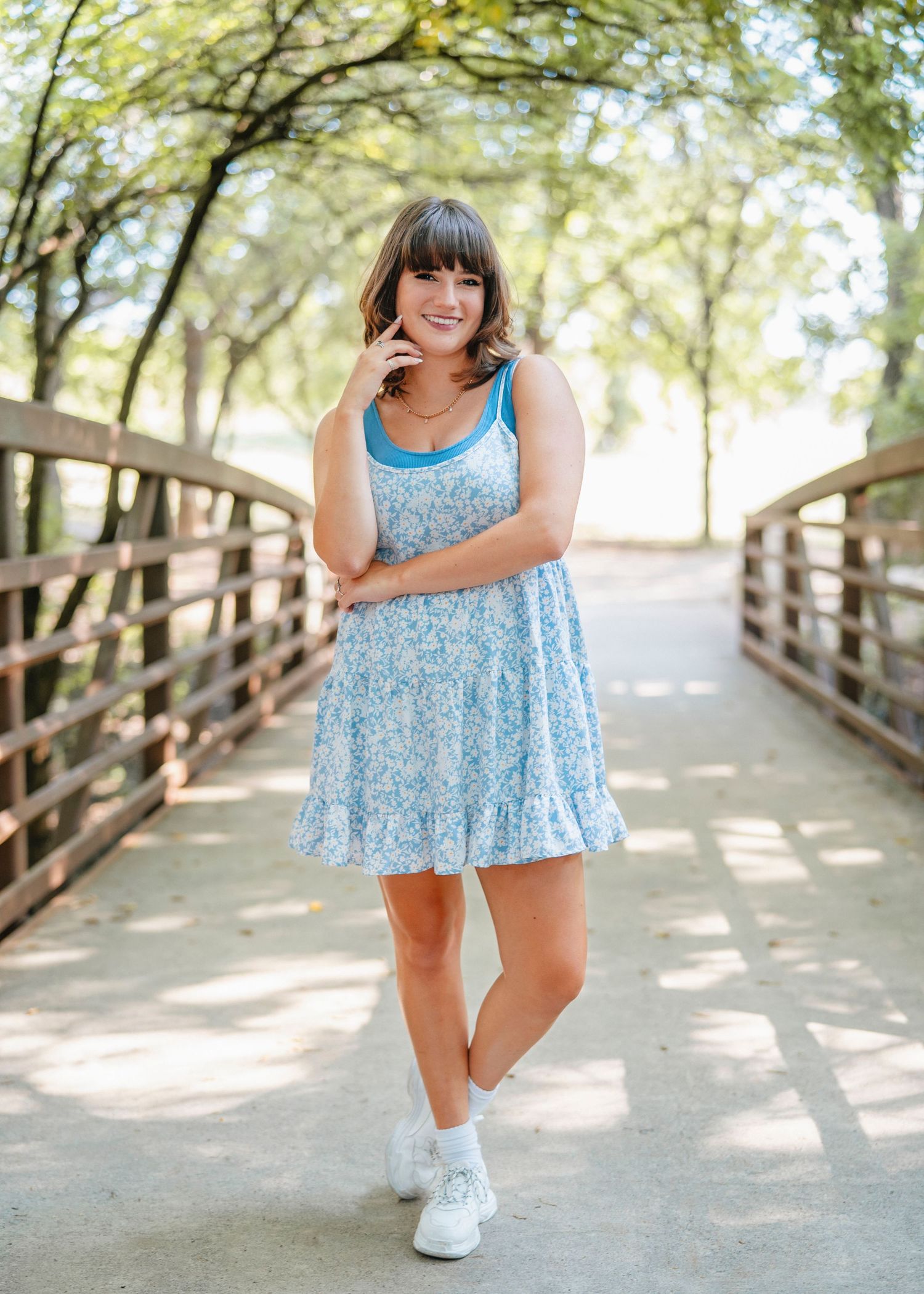Standing on wooden bridge in floral dress with dappled sunlight through trees.