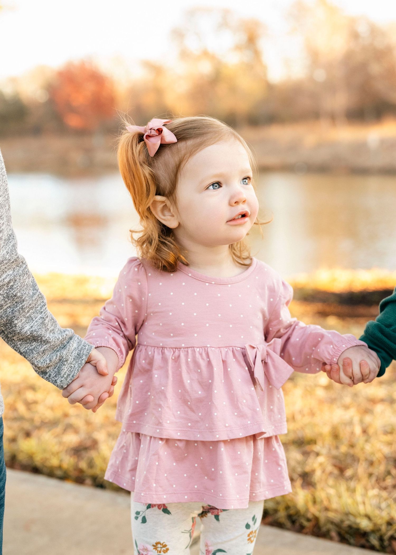 Toddler in pink ruffled dress plays by autumn lake during sunset photo session.
