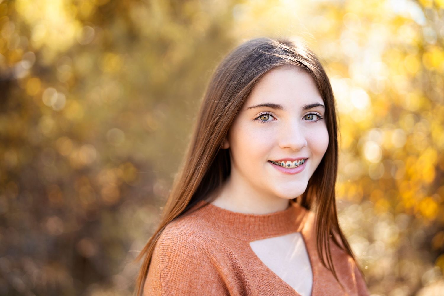 Person in rust-colored sweater posed outdoors with golden autumn trees in background.