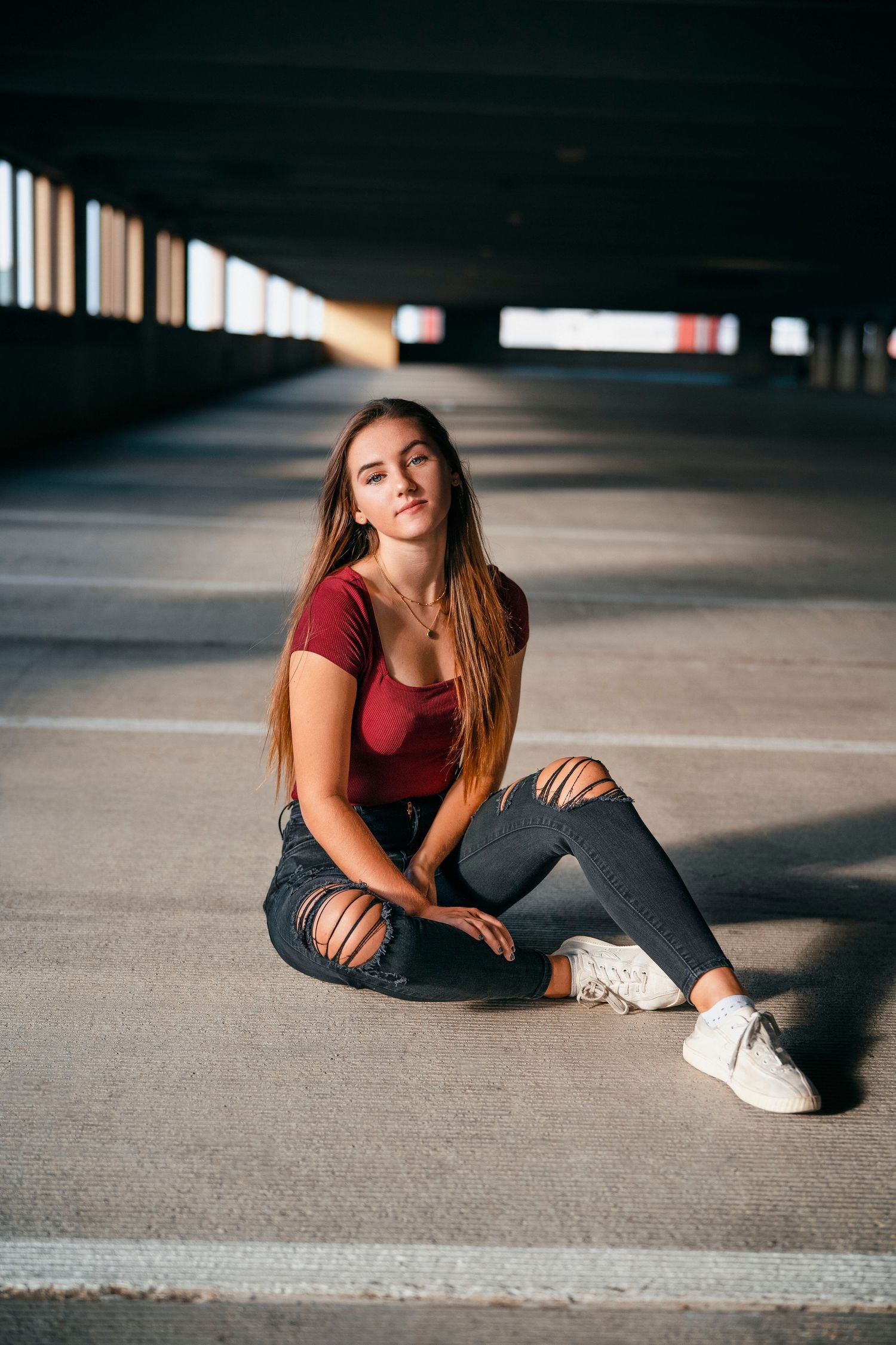 Woman in red top and ripped jeans sitting on floor of urban parking structure.