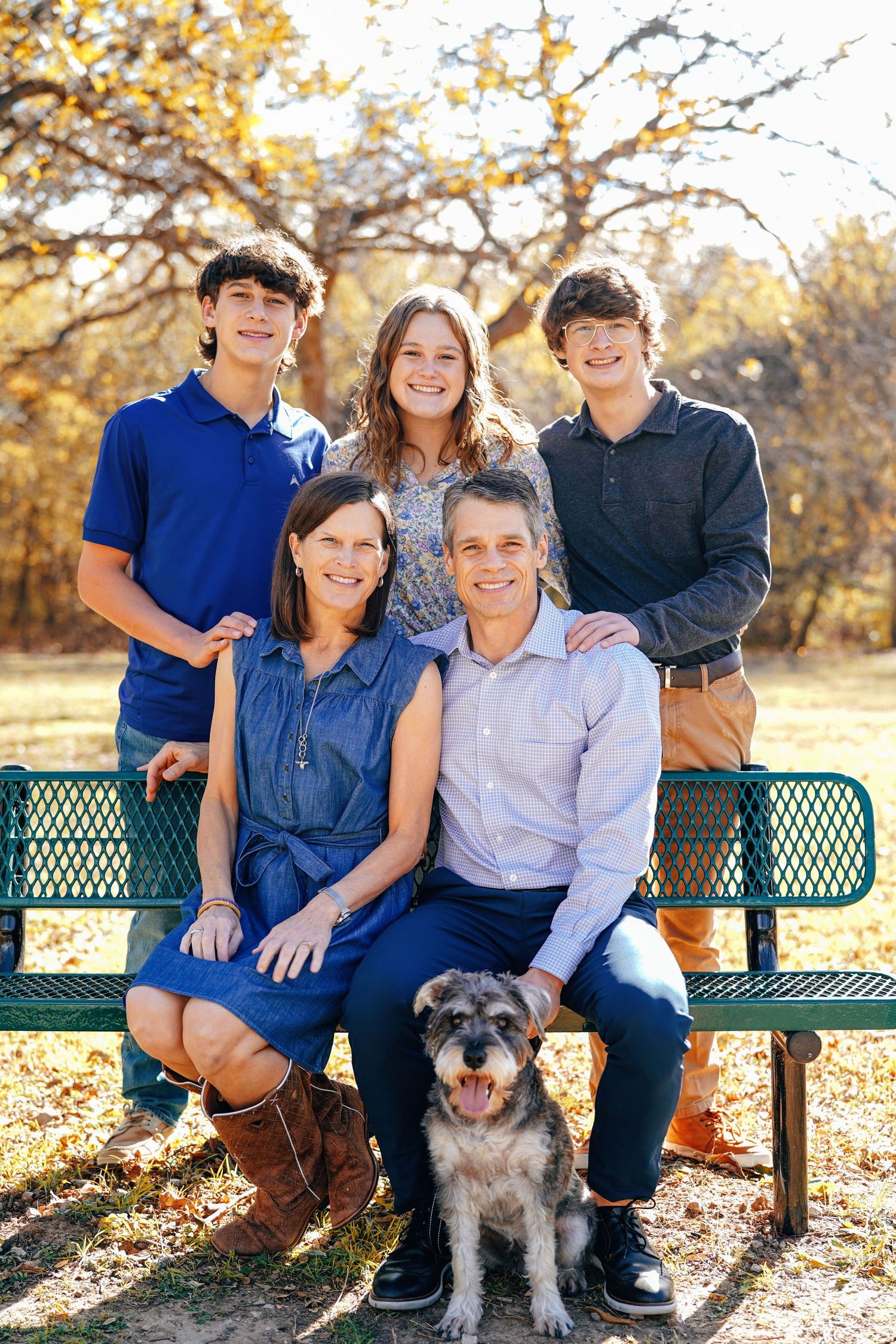 Family sitting on a green park bench with their small scruffy dog during a golden autumn afternoon.