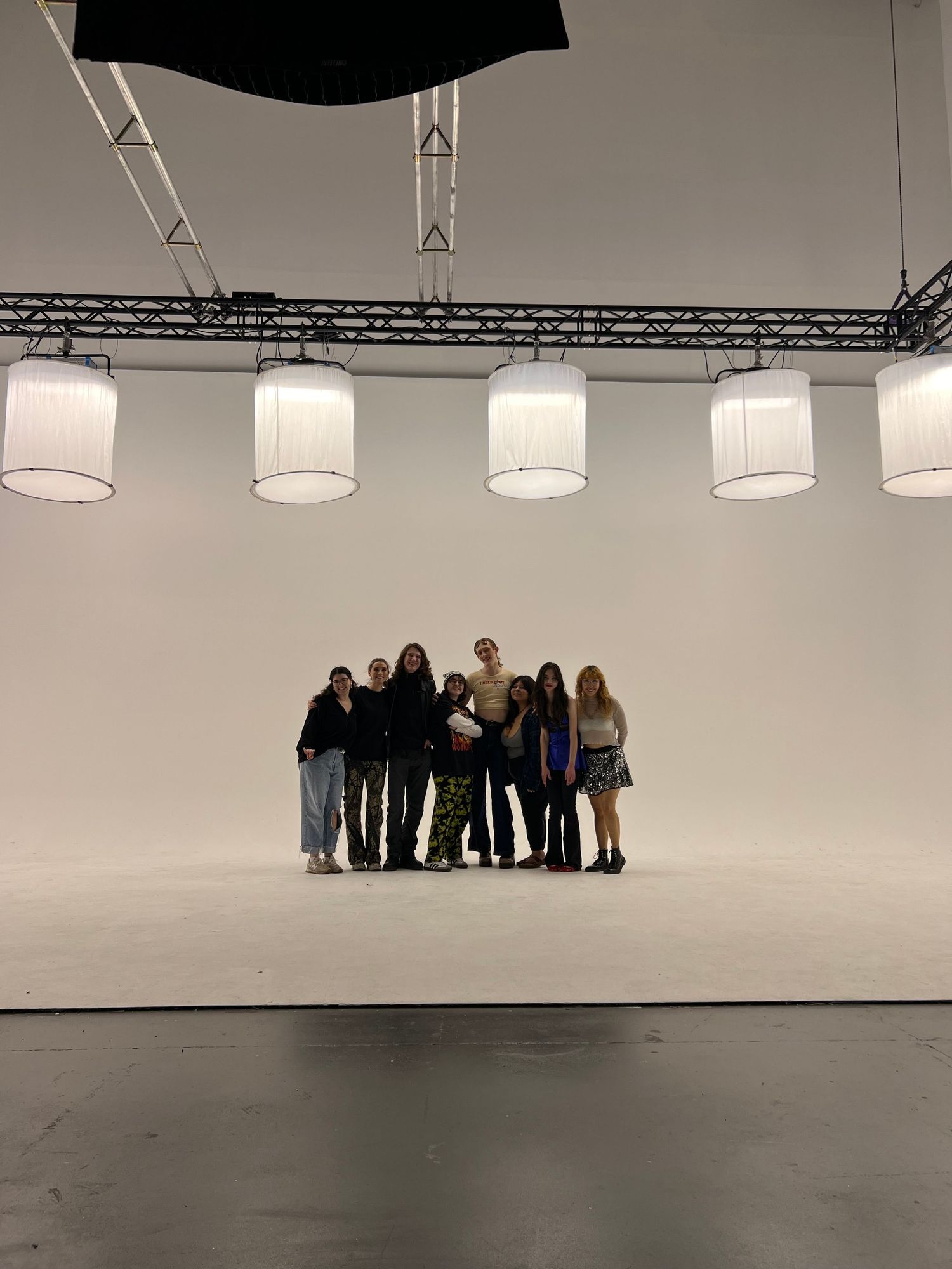A group stands together on a white studio set under hanging cylindrical lighting fixtures.