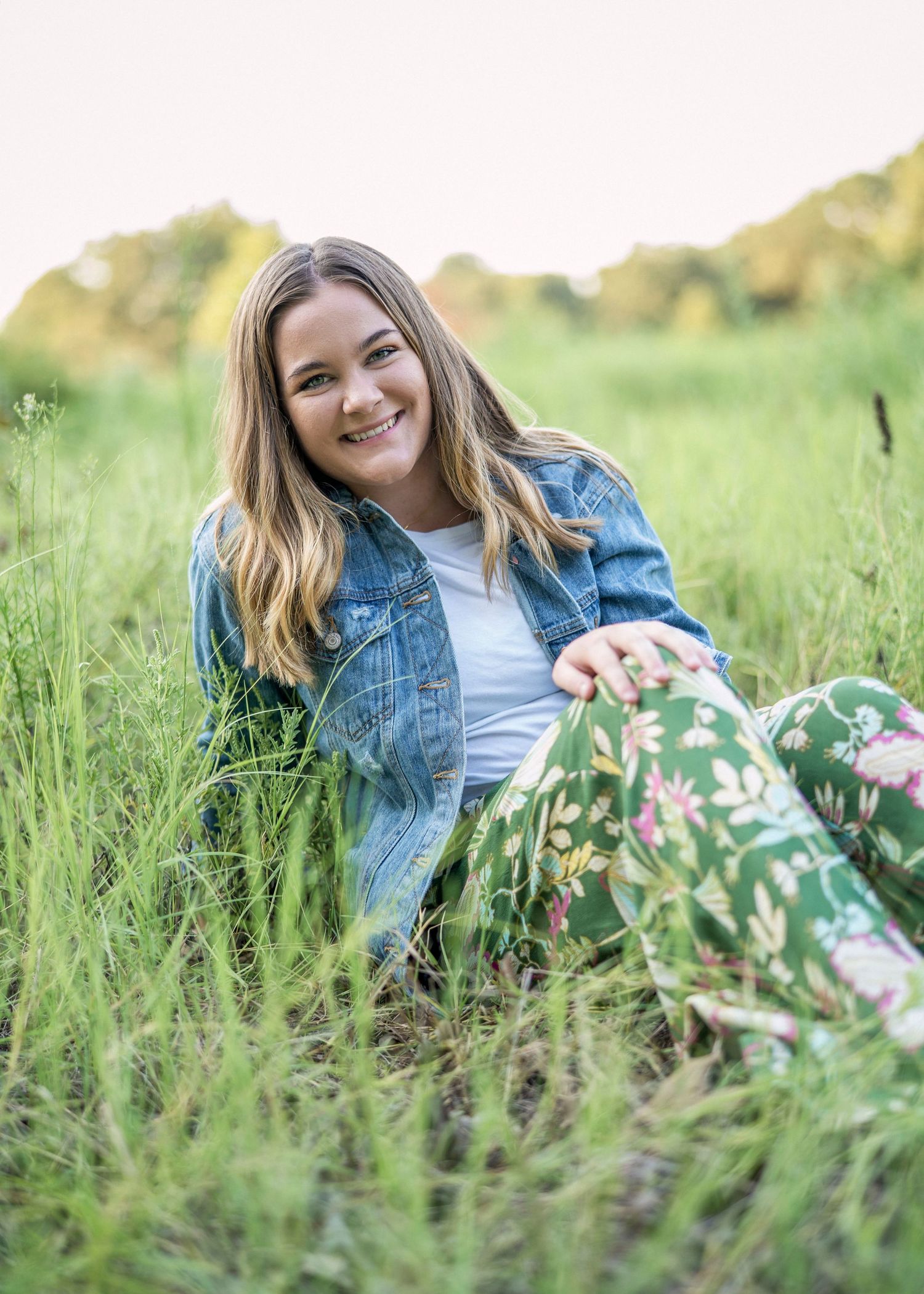 Person relaxing in tall grass wearing denim jacket and floral pants during sunset.
