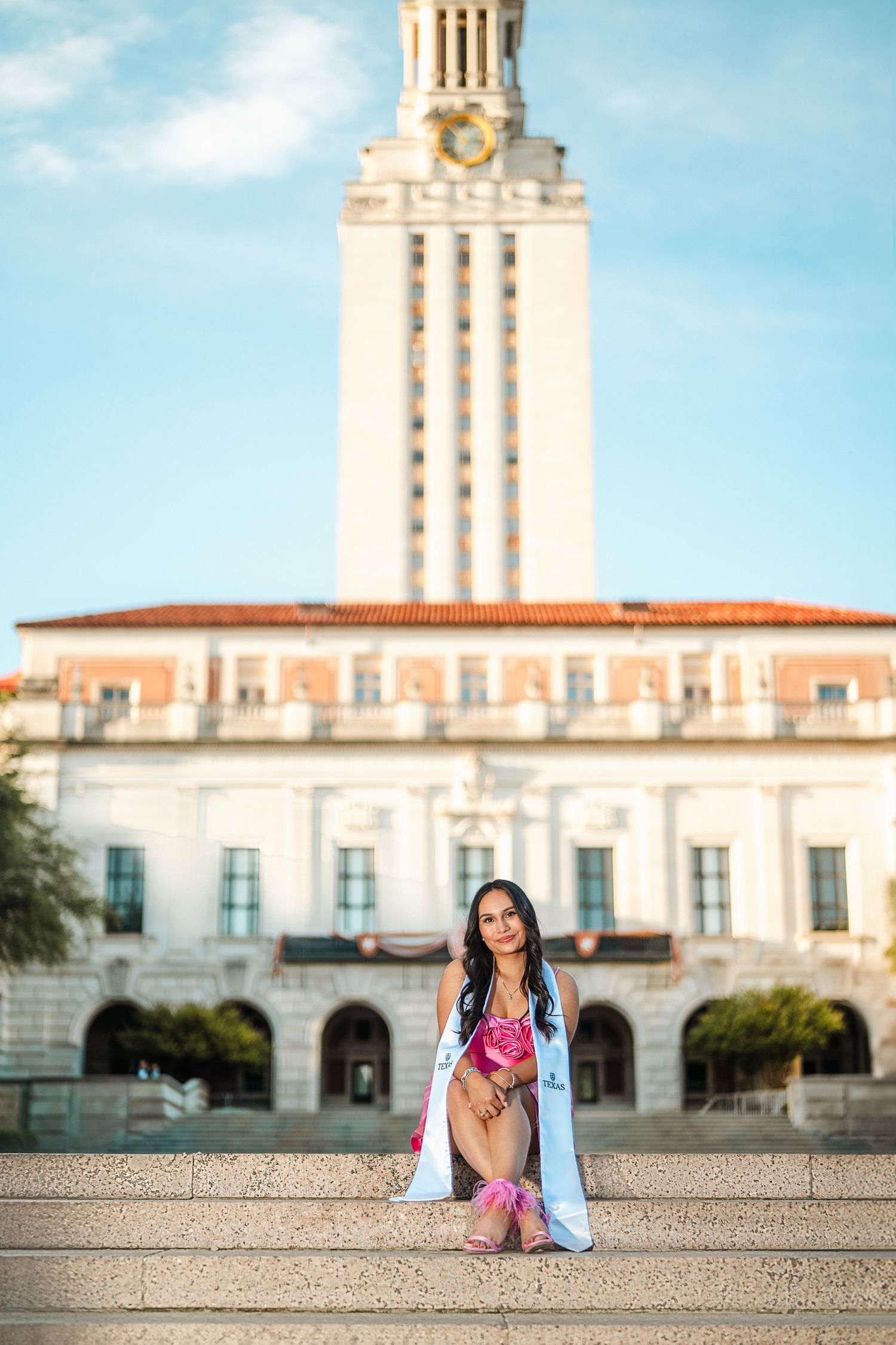 Graduate in blue cap and gown poses on steps of iconic university tower building.