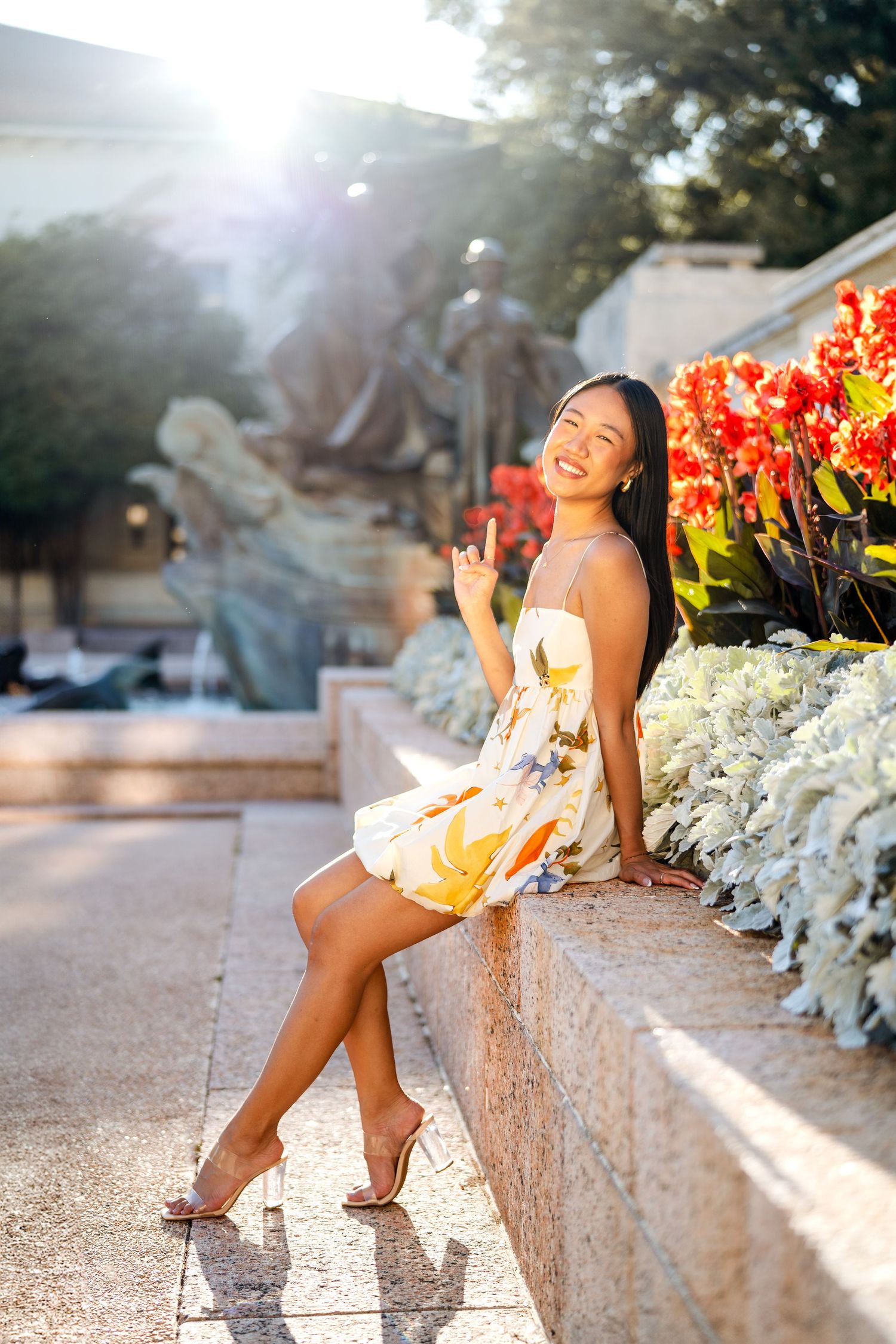 Woman in floral dress sits on stone ledge by garden during golden hour.