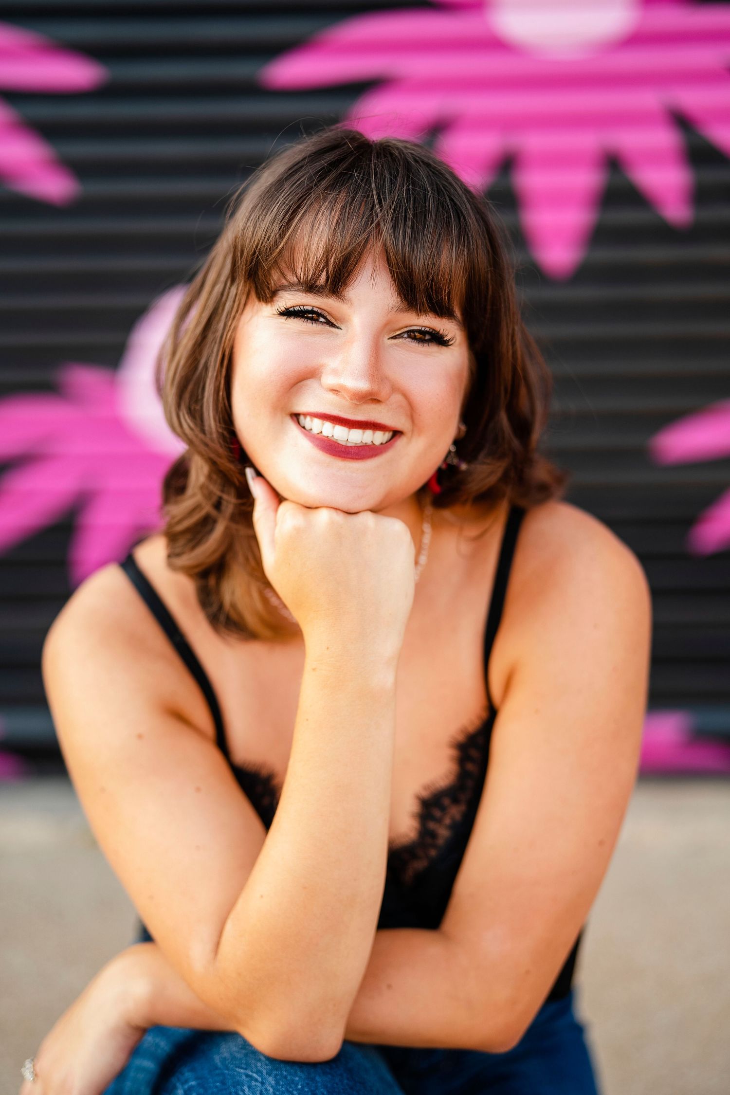 Portrait of someone in a black lacy top smiling against a wall with pink graffiti artwork.