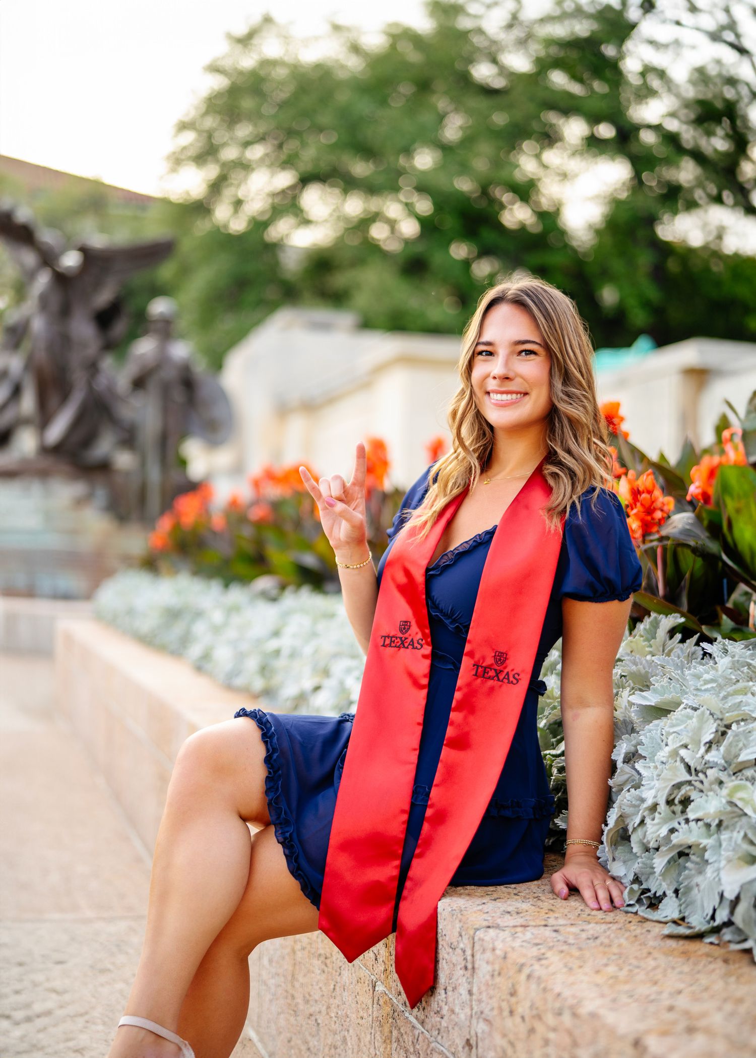 Happy graduate in navy dress makes peace sign while sitting by garden wall with orange flowers.