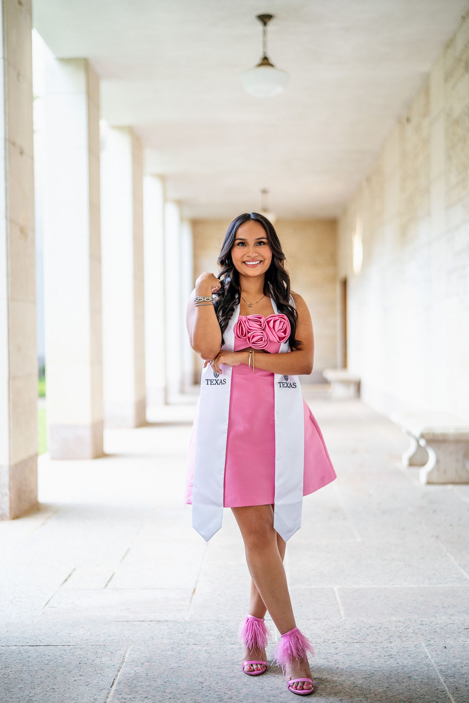 Graduate in pink dress poses in white columned hallway during graduation photoshoot.