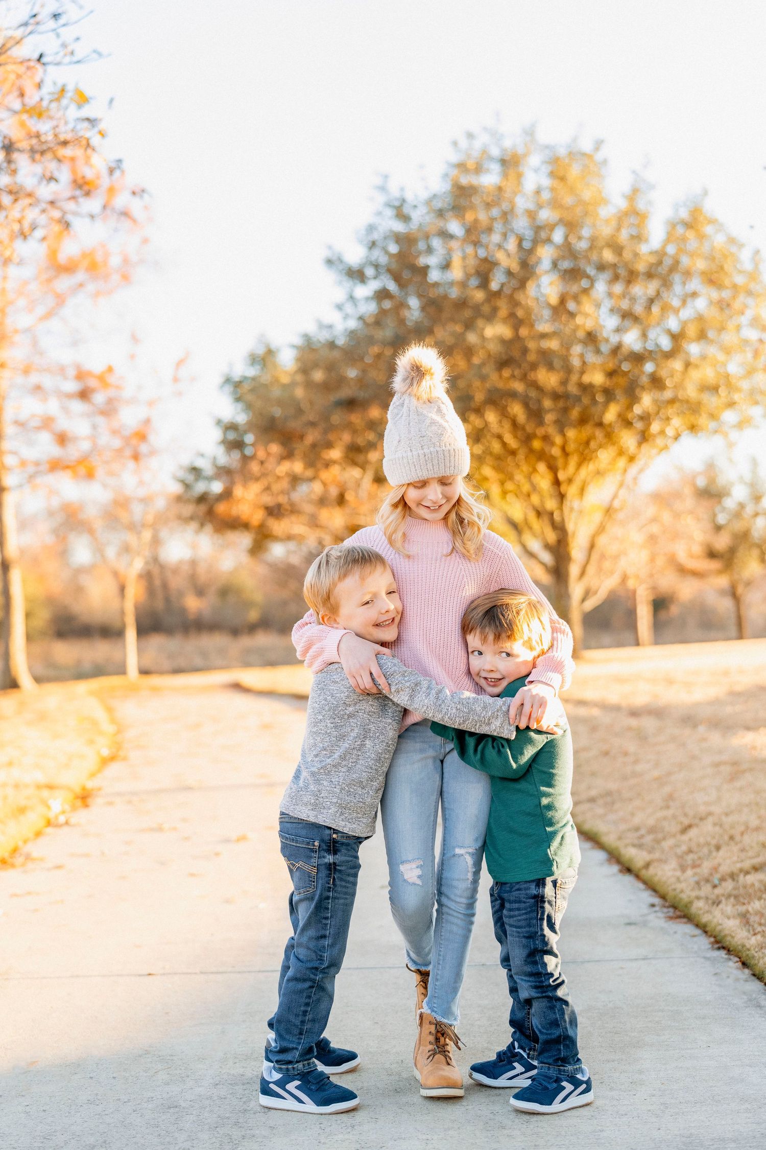 Person in pink sweater and beanie gets hugs from two children on outdoor path at sunset.