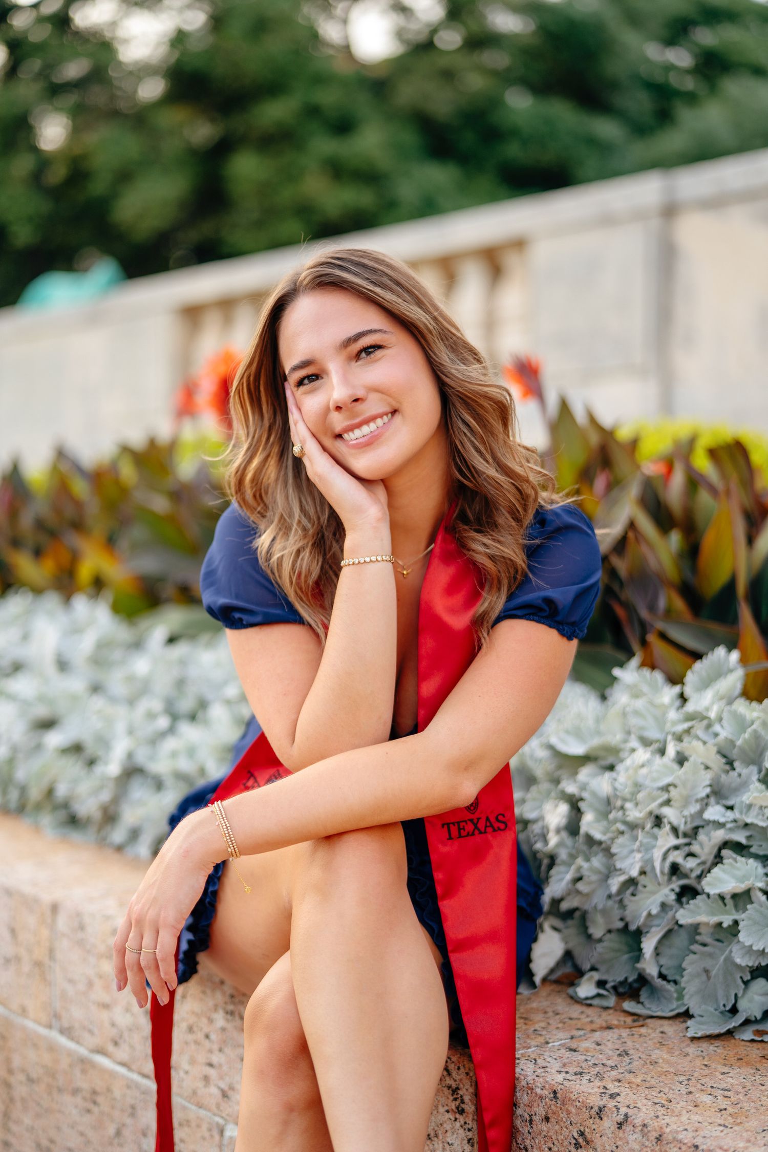 Graduate in navy dress and red stole poses casually by garden wall with dusty miller plants.