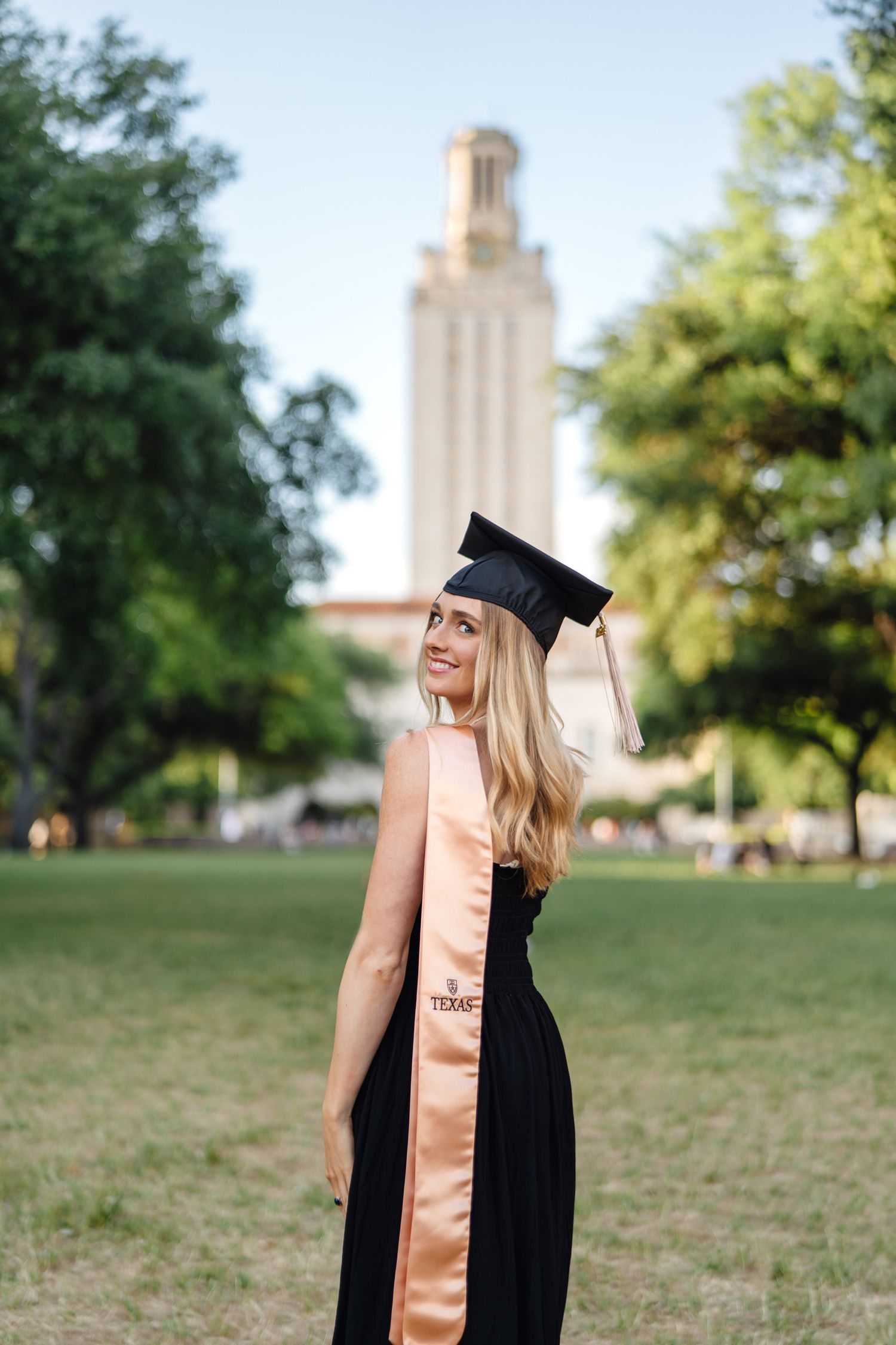 Graduate in black dress and peach sash poses in front of iconic university tower.