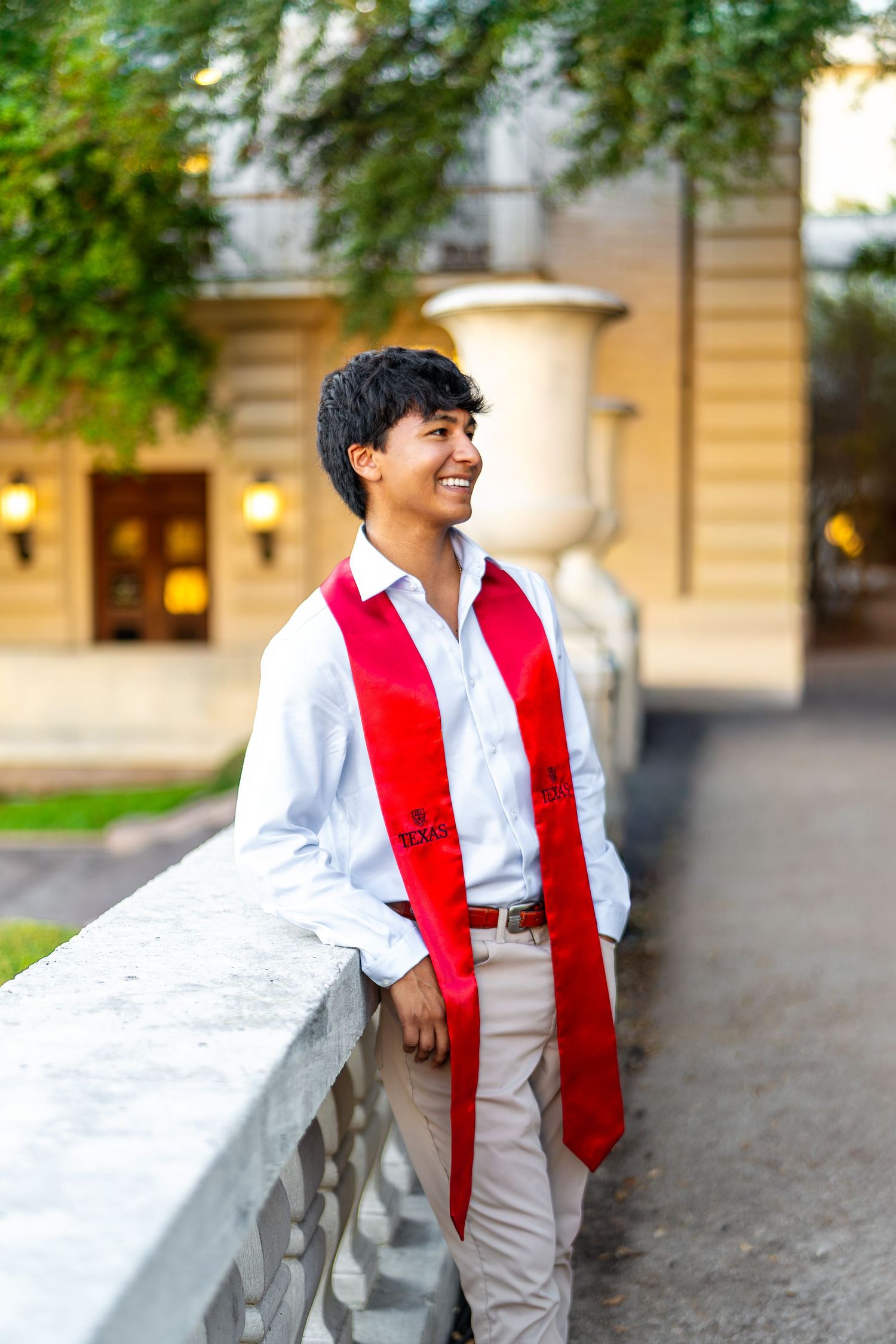 A child in a white shirt and red tie leans against a stone wall while smiling in an outdoor setting.