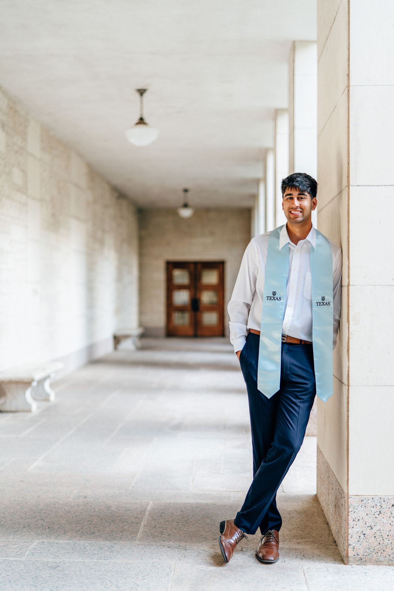 Graduate in light blue stole and dark suit poses confidently in a bright hallway corridor.