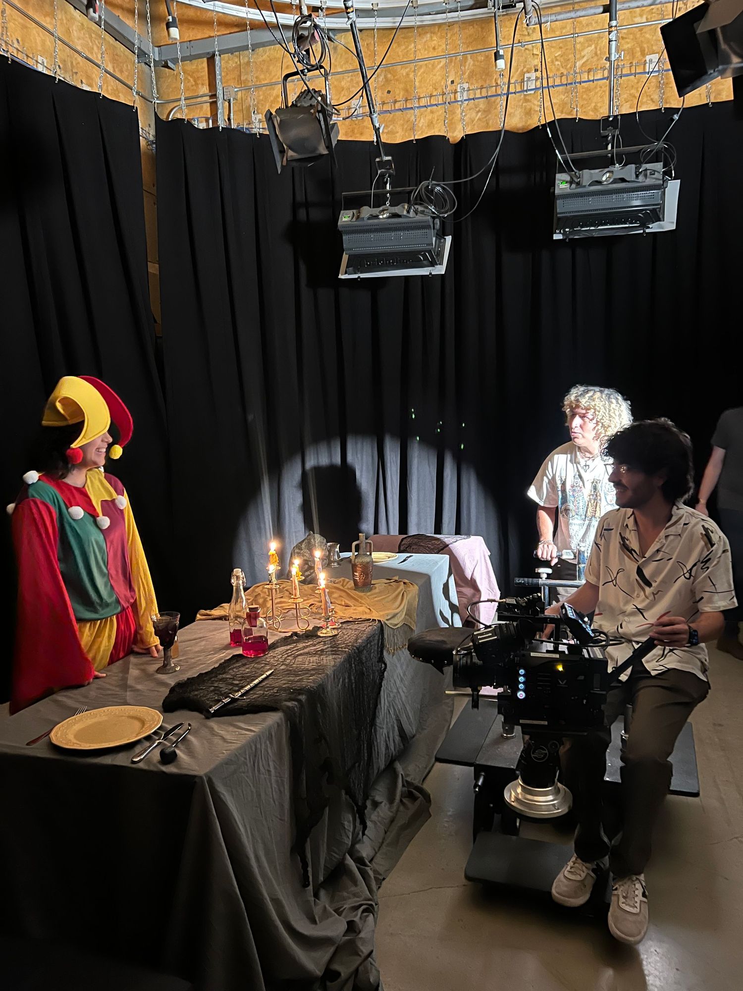 Behind-the-scenes film shoot with camera operator and colorful jester costume on display against black backdrop.