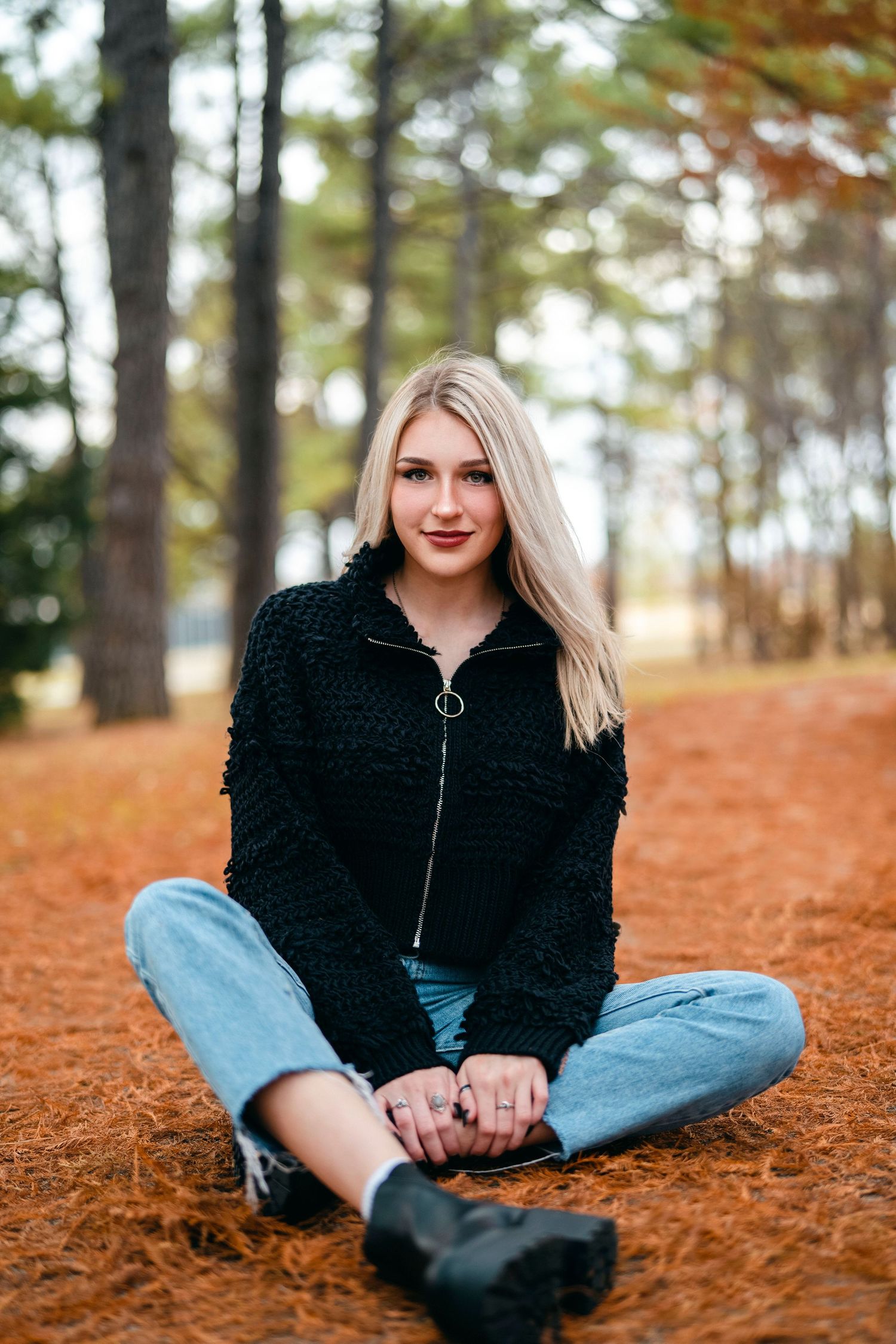 Person in black sweater and jeans sitting on ground covered with fall pine needles.