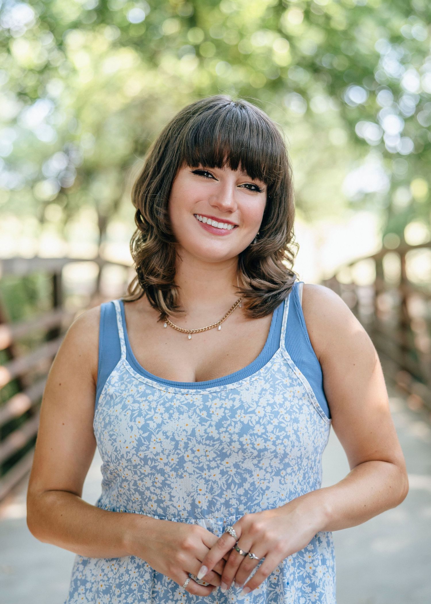 Portrait of someone in a blue floral tank top smiling outdoors with soft bokeh background.