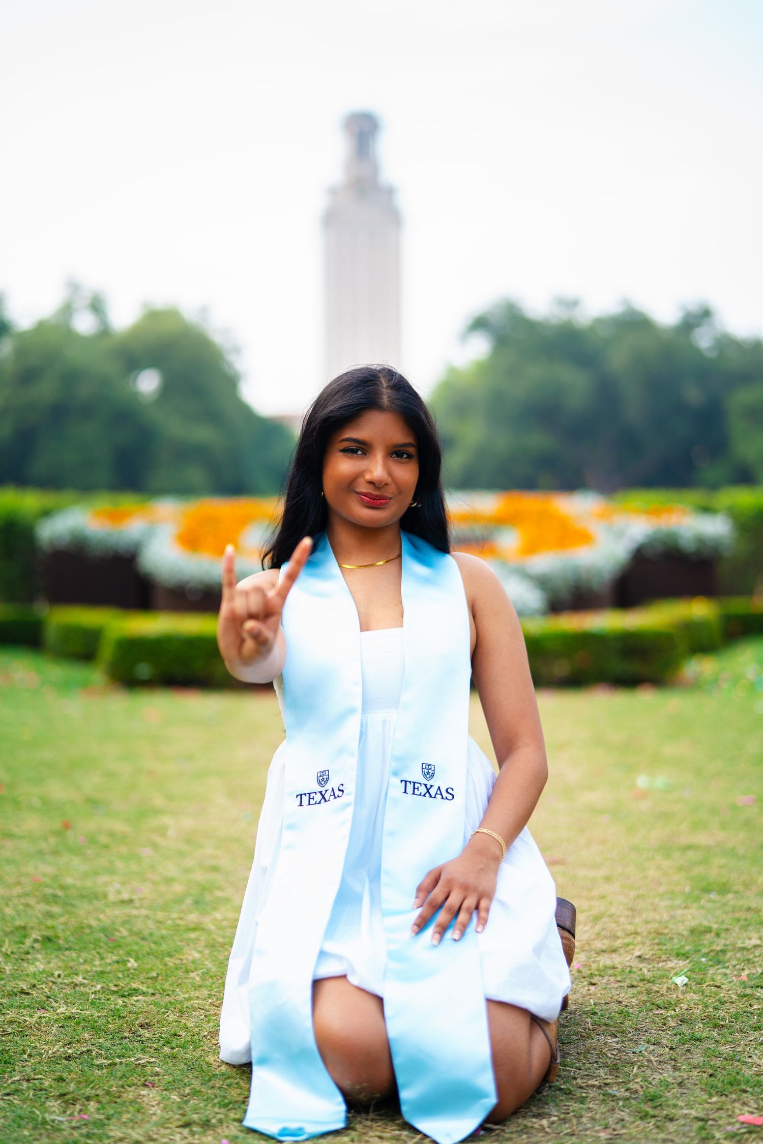 Graduate in white dress and blue stole kneels for photo with UT Tower backdrop.