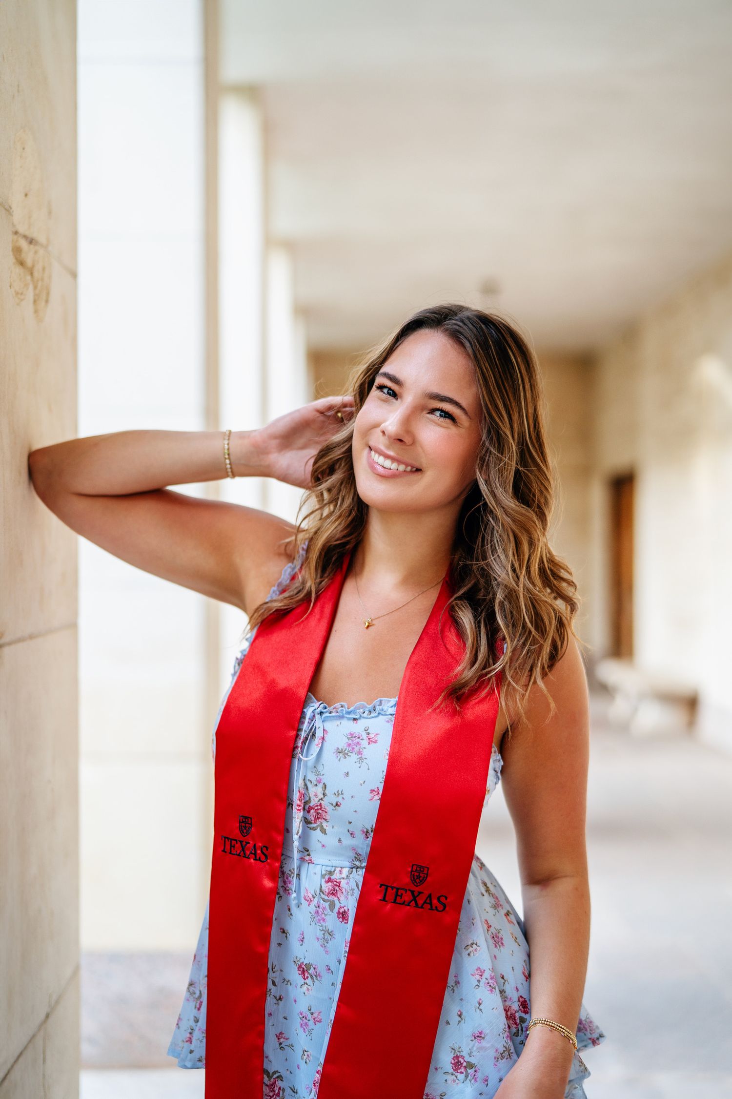 Graduate in floral dress and red stole smiling in outdoor corridor.
