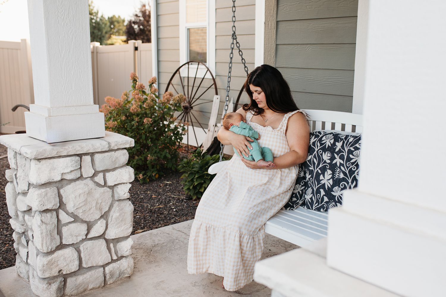 A person in a white dress sits on a front porch with stone columns holding a infant - lifestyle in-home newborn photos Boise