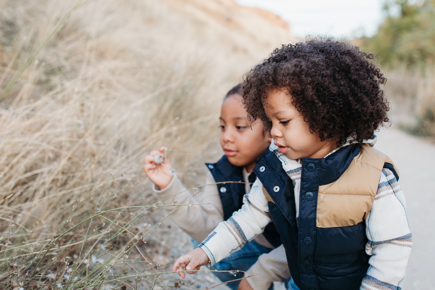 Two children in puffer vests explore nature together while examining something interesting in the grass. - lifestyle family photos in Boise