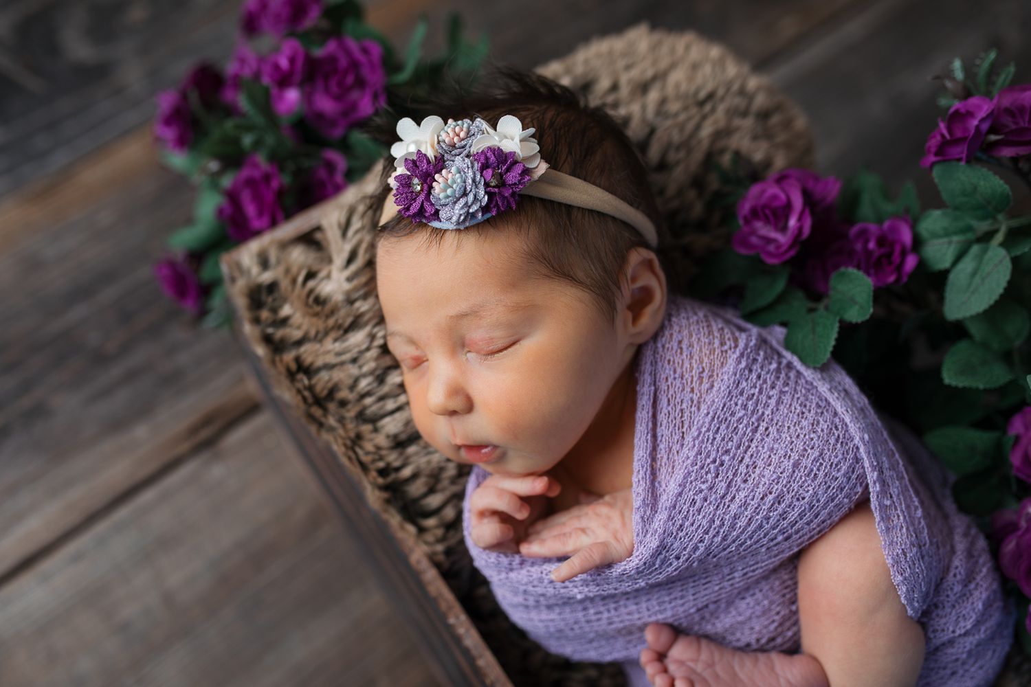 A sleeping newborn in a purple wrap and headband poses peacefully against a rustic wooden backdrop with purple flowers. studio newborn