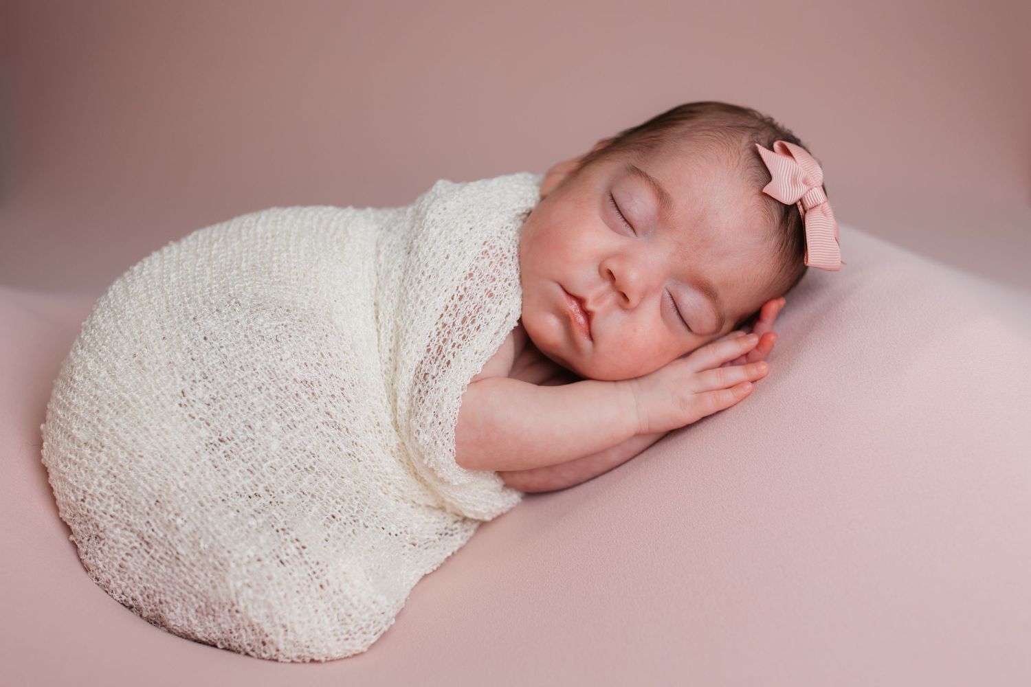 Sleeping newborn baby girl posed in a cream wrap with a pink bow during her studio newborn session