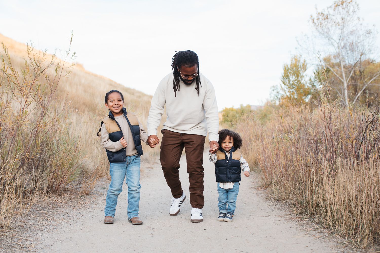 Three people walking together on a dirt path surrounded by tall dried grass and autumn foliage - lifestyle family photos in Boise