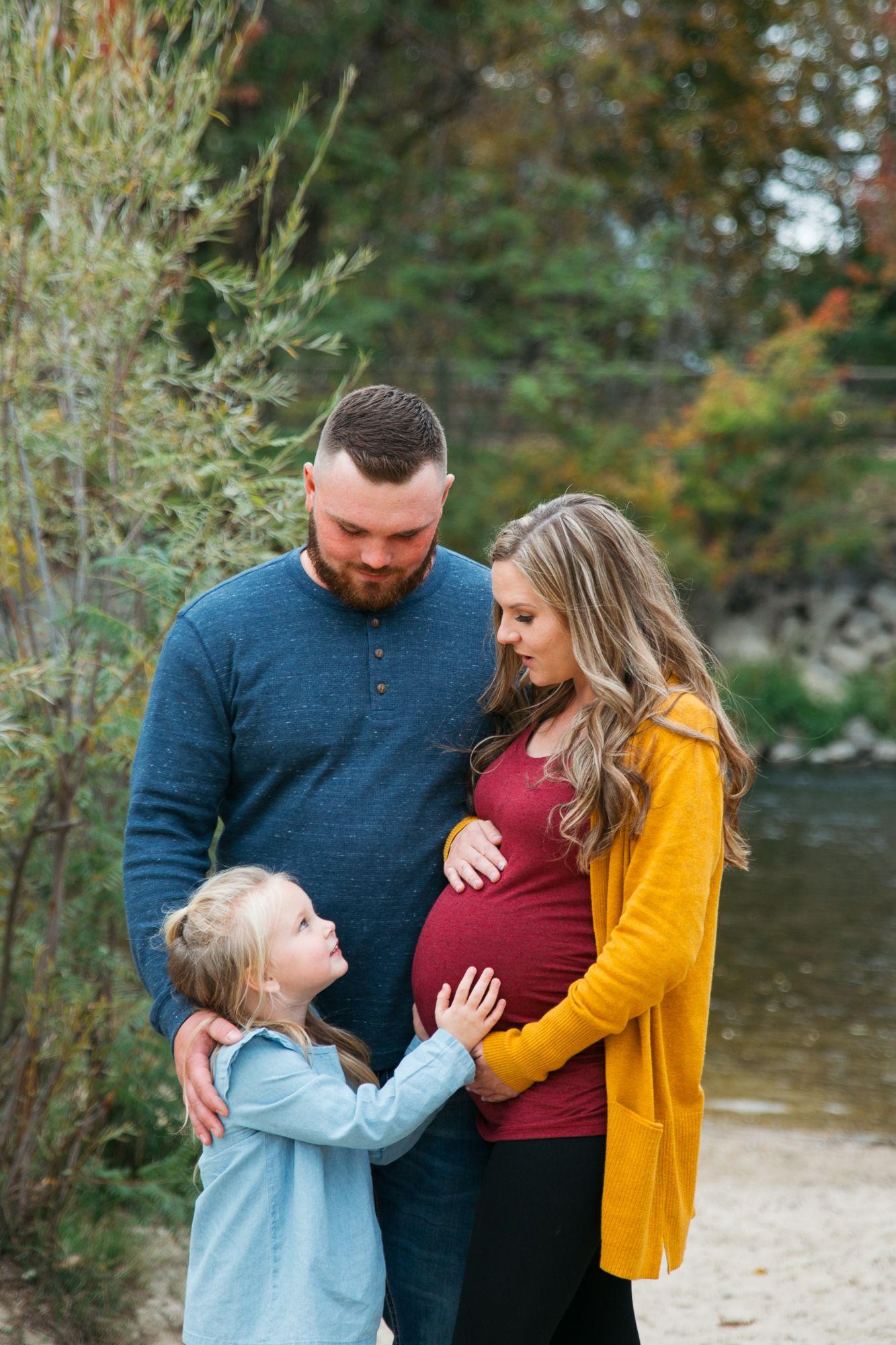 A family of three shares tender moments by a river while wearing fall-colored clothing during an outdoor photo session - Maternity Eagle ID