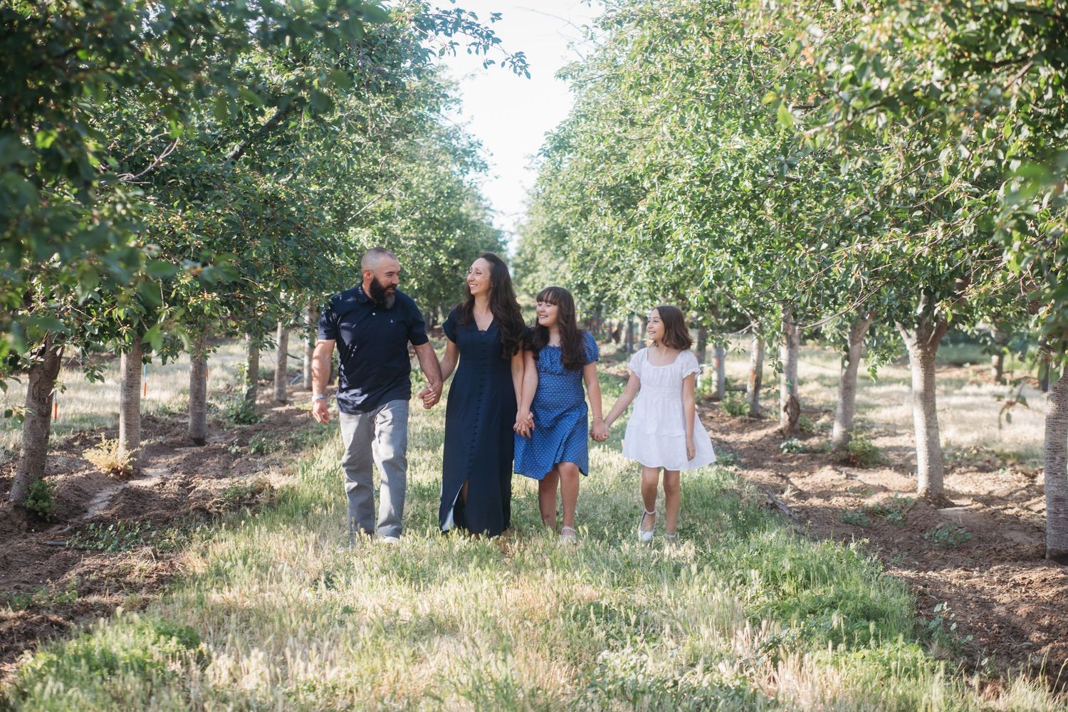 Late spring family session walking through a cherry orchard in Boise, Idaho with parents and two daughters holding hands and smiling