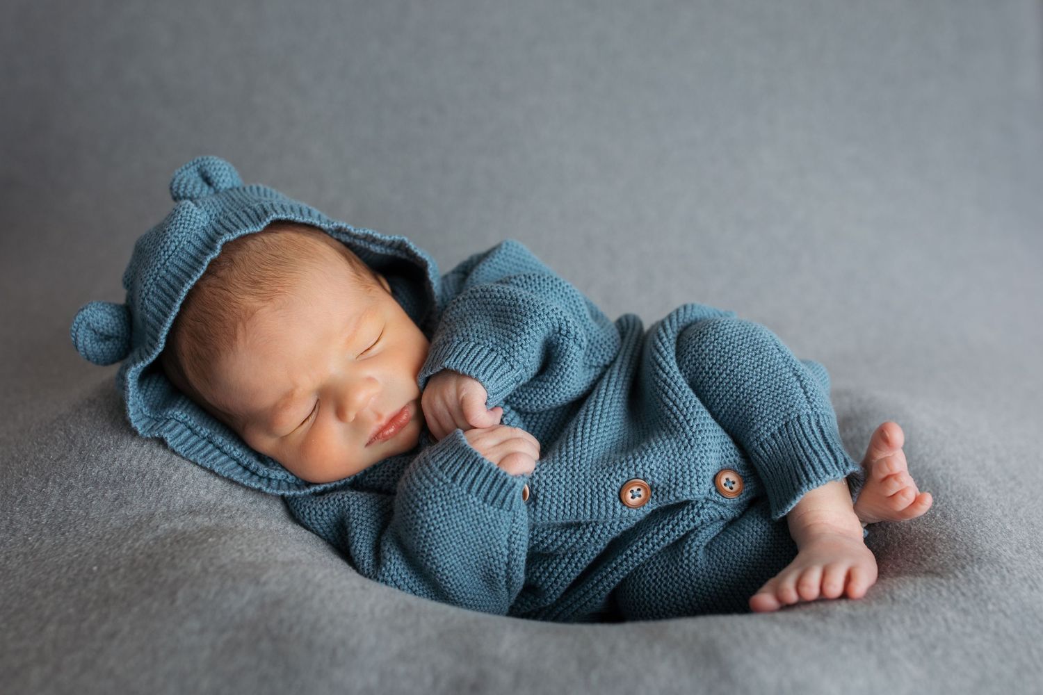 Full-body studio portrait of newborn baby boy sleeping in blue knit outfit on soft gray blanket