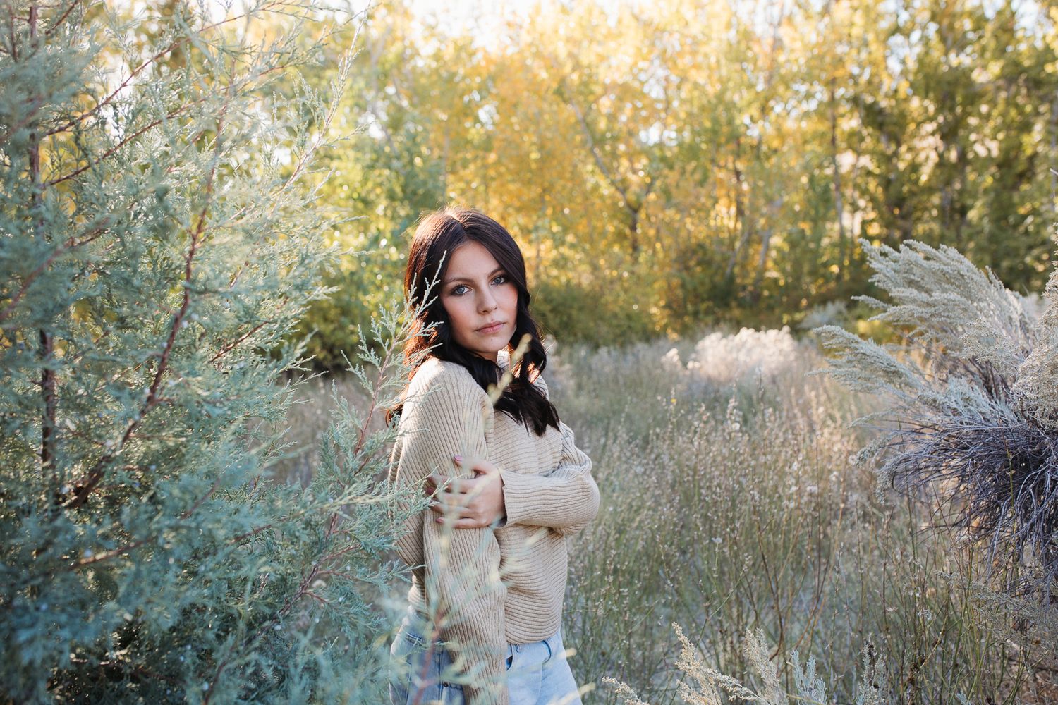 A person in a beige coat poses outdoors among autumn trees and natural scenery during a photoshoot. Boise foothills family photography