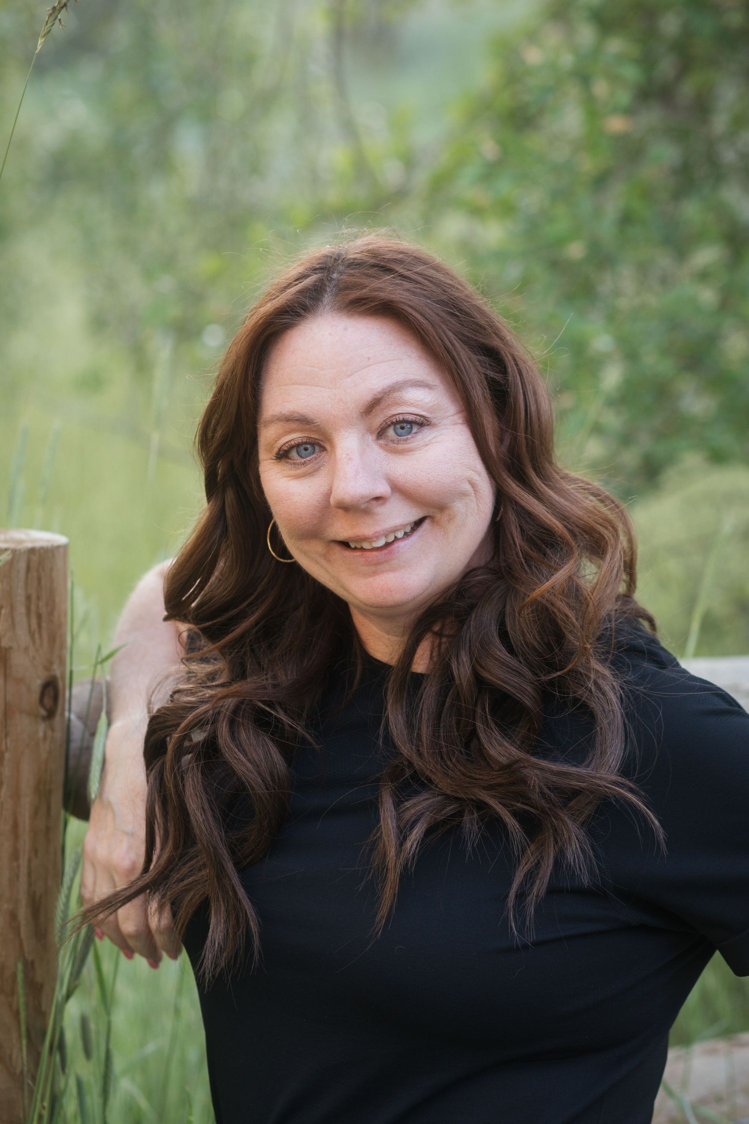Smiling woman with long brown wavy hair in black top leaning against wooden fence outdoors.
